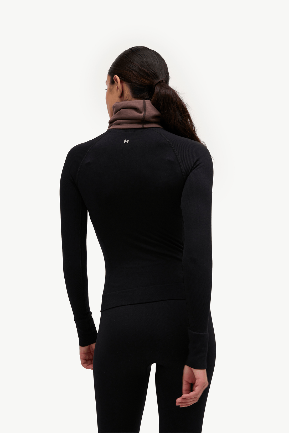 A woman shown from the back wearing a black long-sleeve athletic top with a brown neck gaiter and black leggings against a plain white studio background.