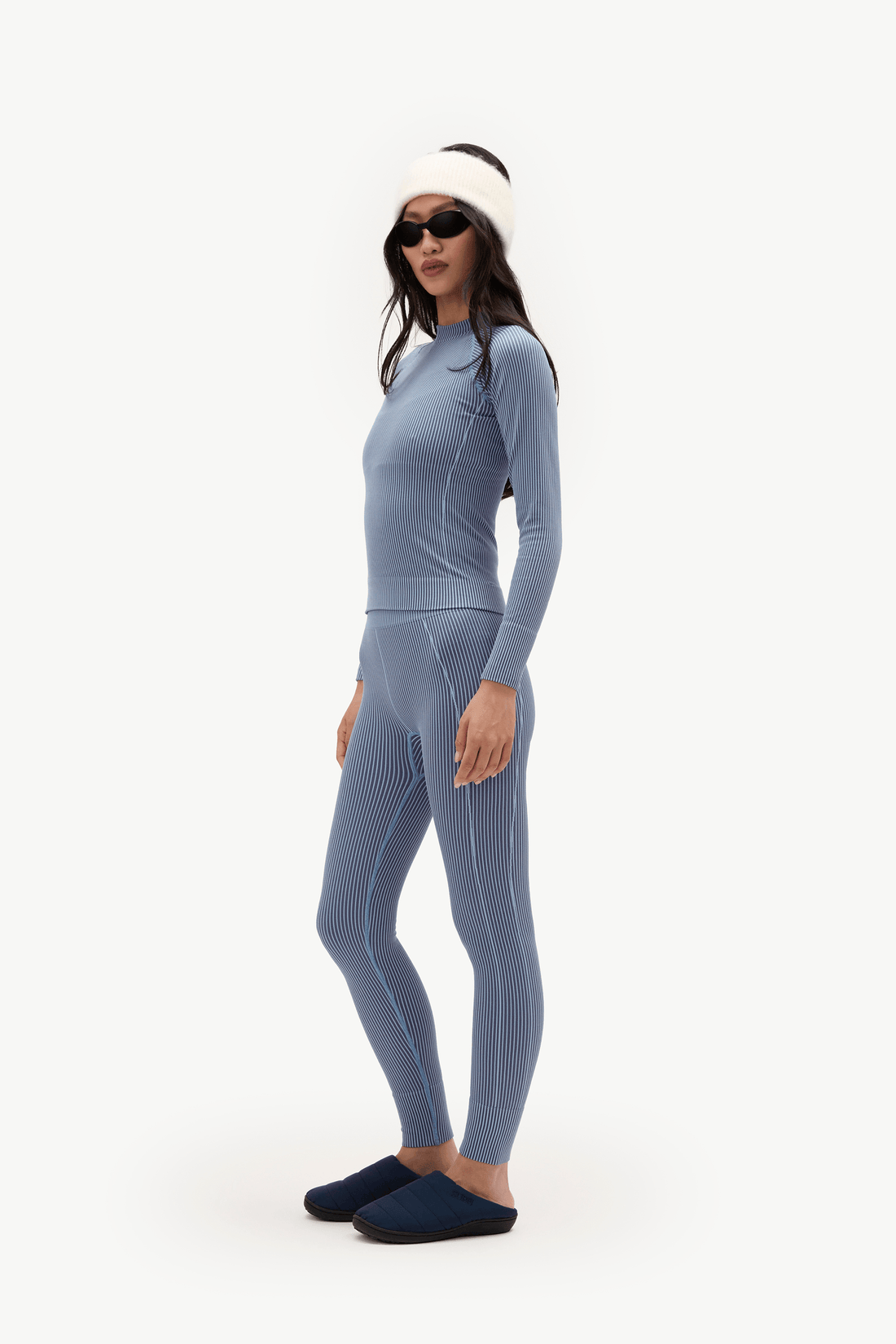 A woman modeling a matching ribbed knit two-piece set in a dusty blue-gray color, consisting of a long-sleeve top and leggings, styled with a white beanie, black sunglasses, and dark slip-on shoes against a plain white studio background.
