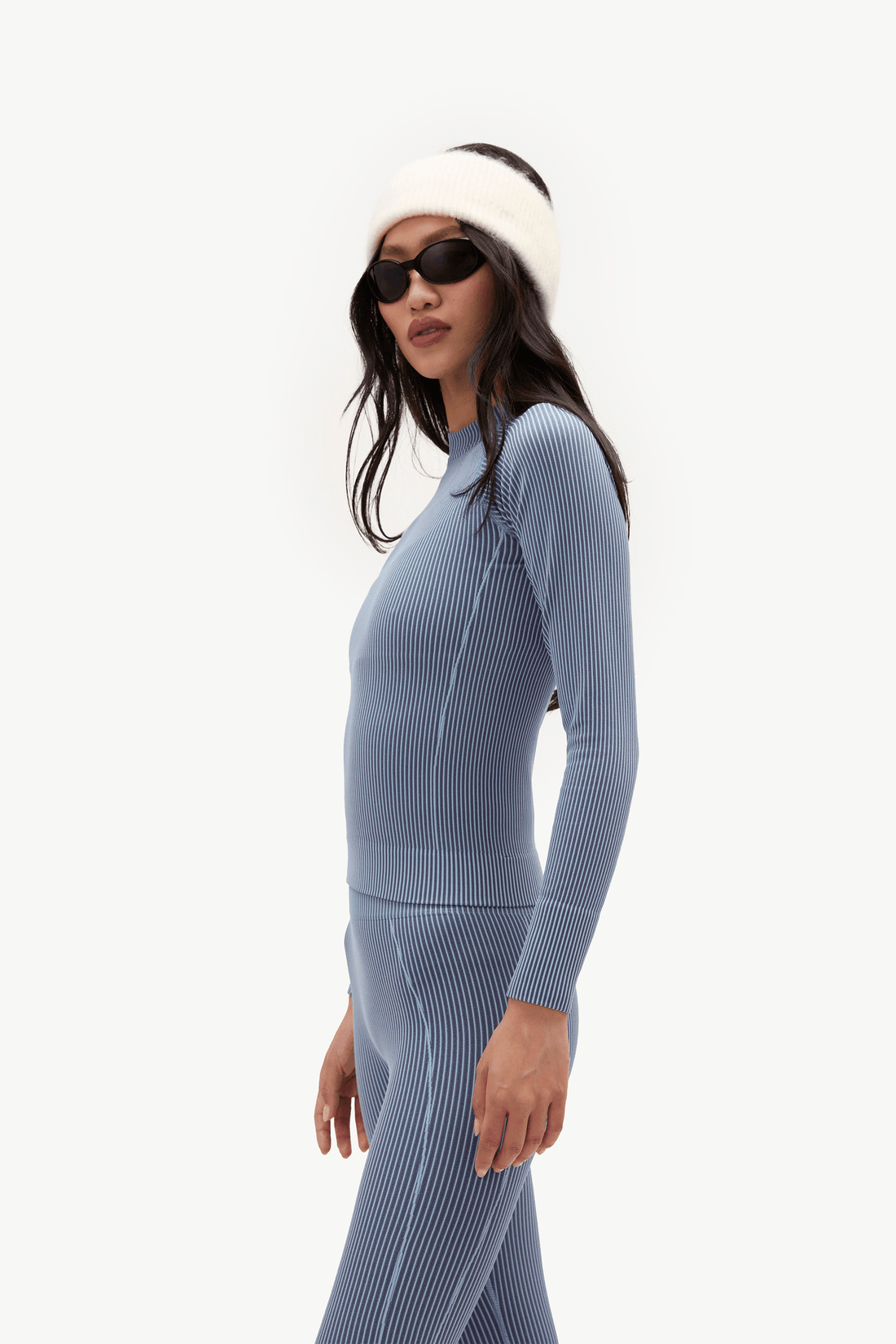 A woman in side profile wearing a matching ribbed knit two-piece outfit in dusty blue, including a mock-neck long-sleeve top and high-waisted leggings, styled with a white fuzzy beanie and black sunglasses against a plain white background.