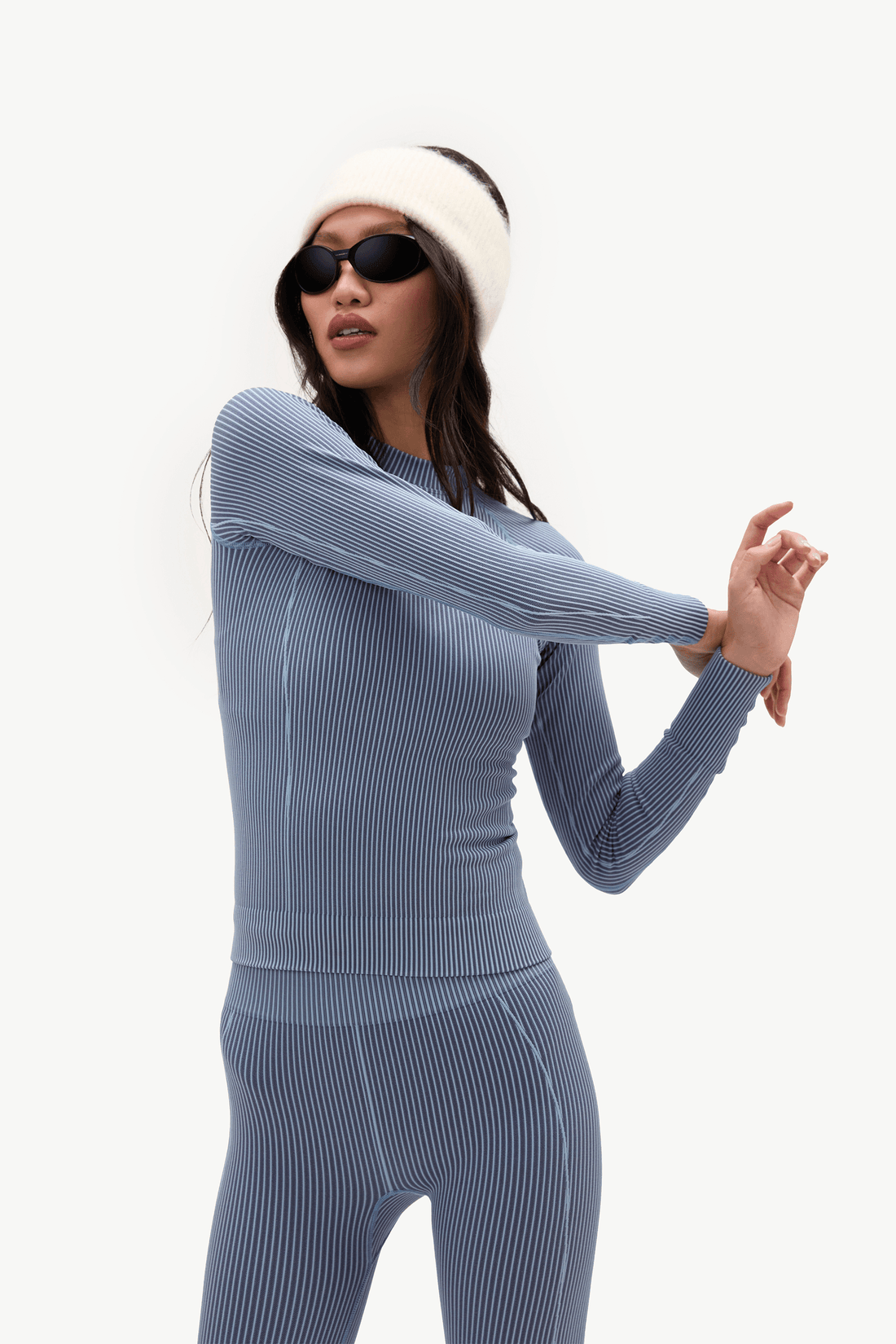 A woman modeling a matching ribbed knit two-piece outfit in a dusty blue-gray color, consisting of a long-sleeve top and leggings, styled with a white beanie and black sunglasses against a plain white background.