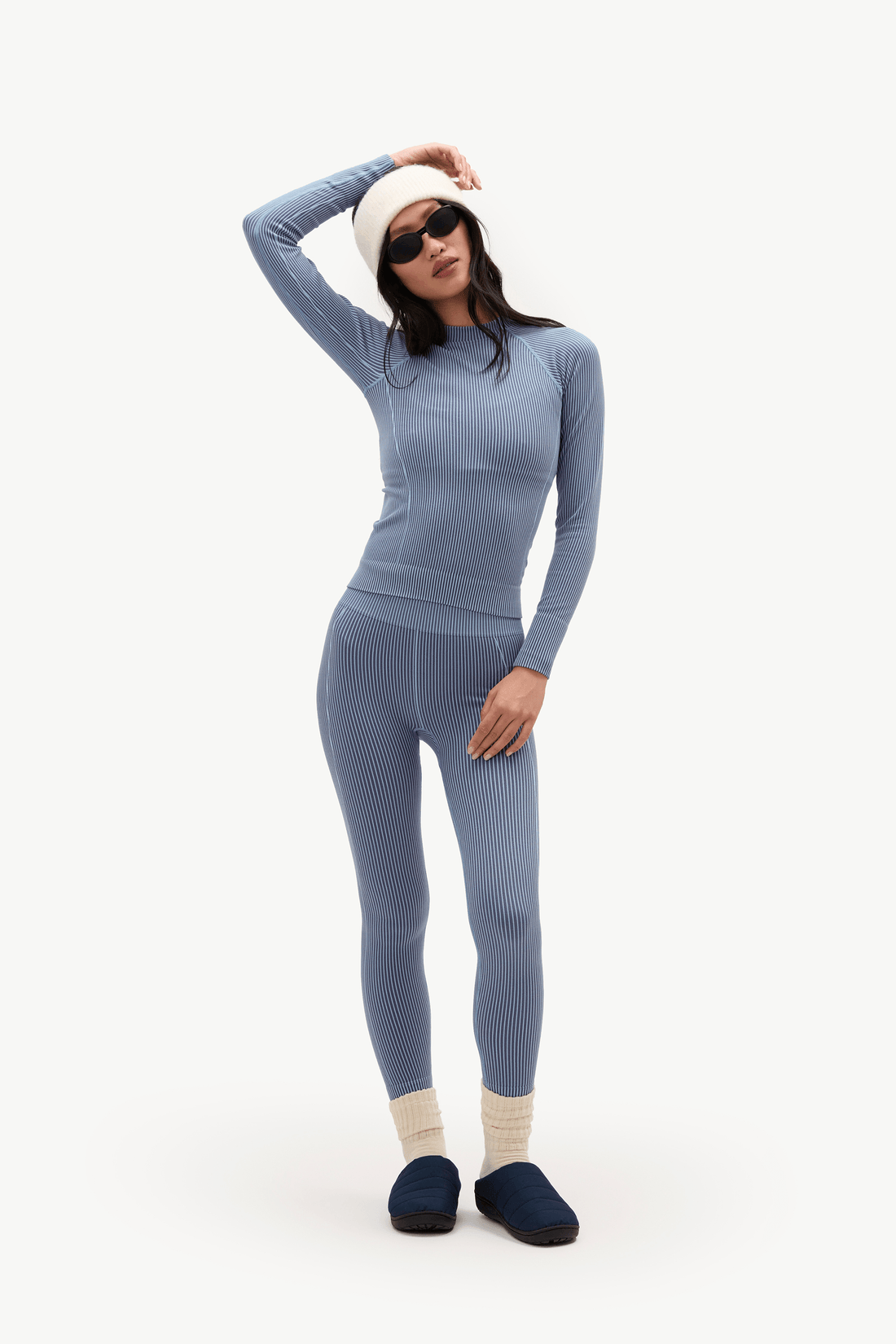 A woman modeling a matching two-piece athletic set in a dusty blue-gray color, consisting of a long-sleeve top and high-waisted leggings. She is also wearing a white beanie, white socks, and dark blue slippers against a plain white studio background.