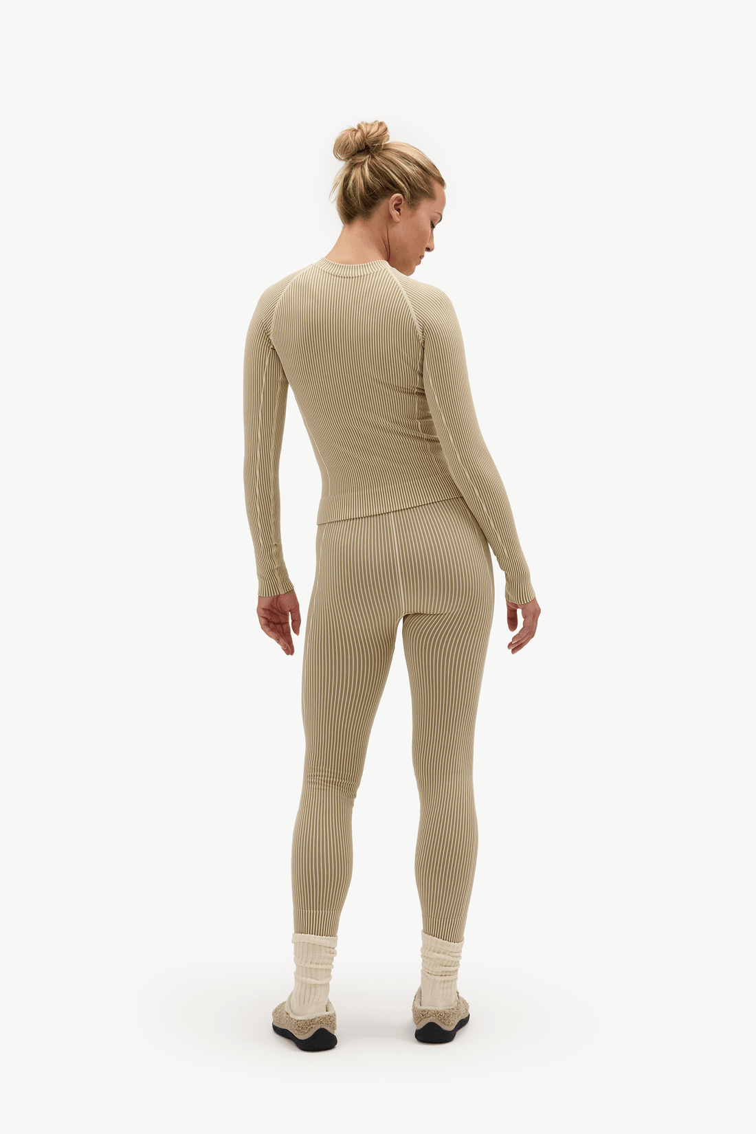 A woman shown from the back wearing a matching ribbed knit one-piece long-sleeve top and leggings set in a light beige color, paired with cream socks and black shoes, against a plain white studio background.