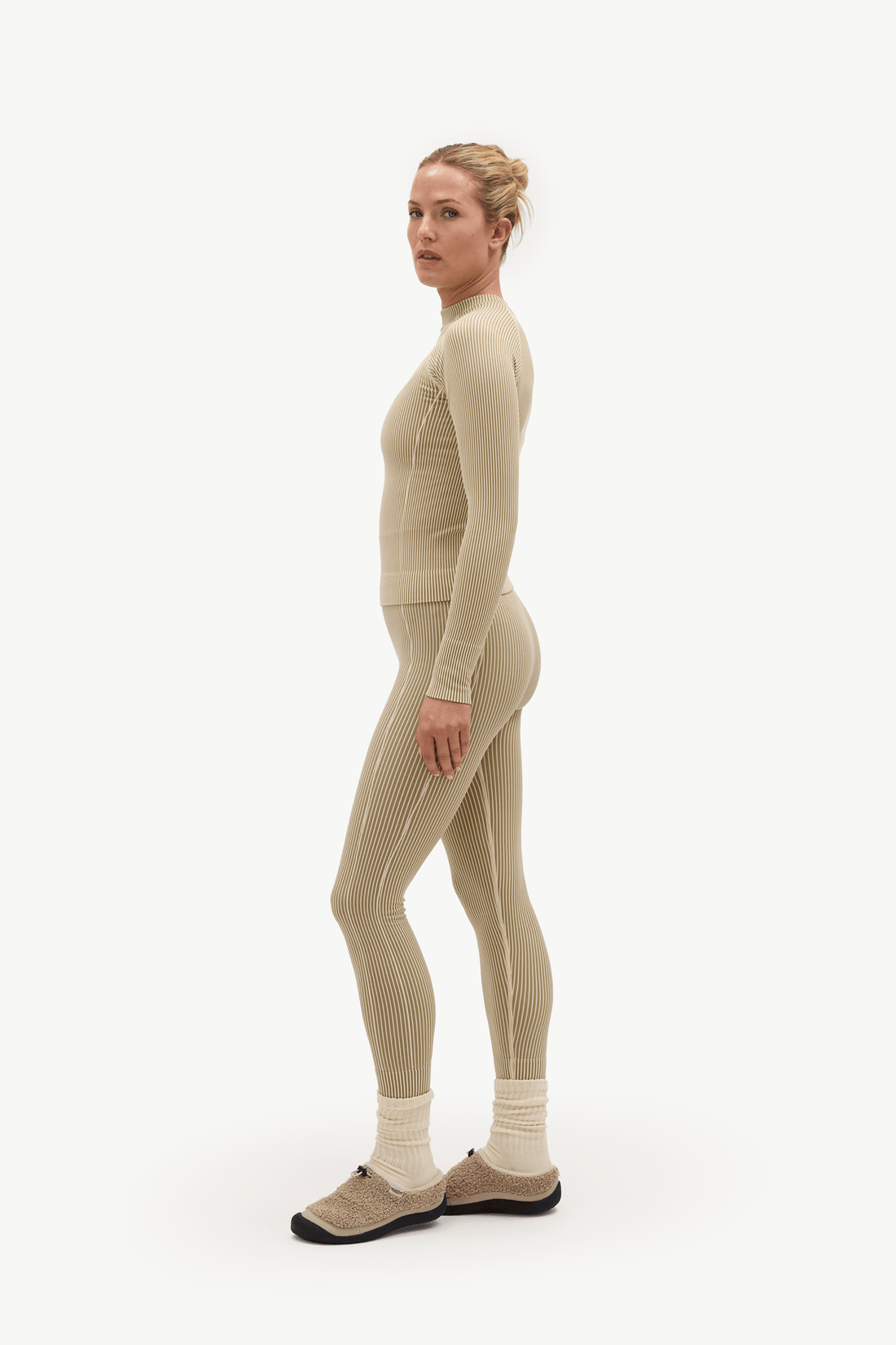 A woman stands in profile wearing a matching ribbed knit one-piece outfit in a light warm beige or oatmeal tone, with white socks and tan shoes, against a plain light gray studio background.
