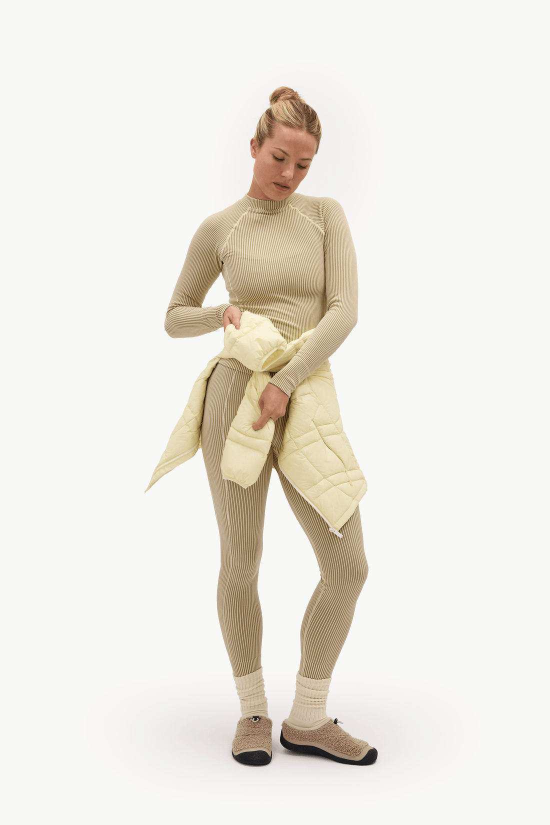A woman modeling a beige ribbed long-sleeve top and matching leggings, holding a pale yellow quilted puffer jacket against a plain white studio background.