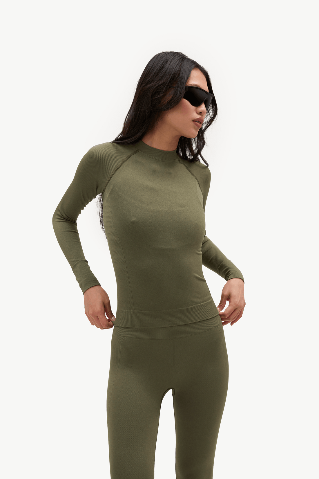 A woman modeling a matching olive green long-sleeve athletic top and leggings set against a plain white background.