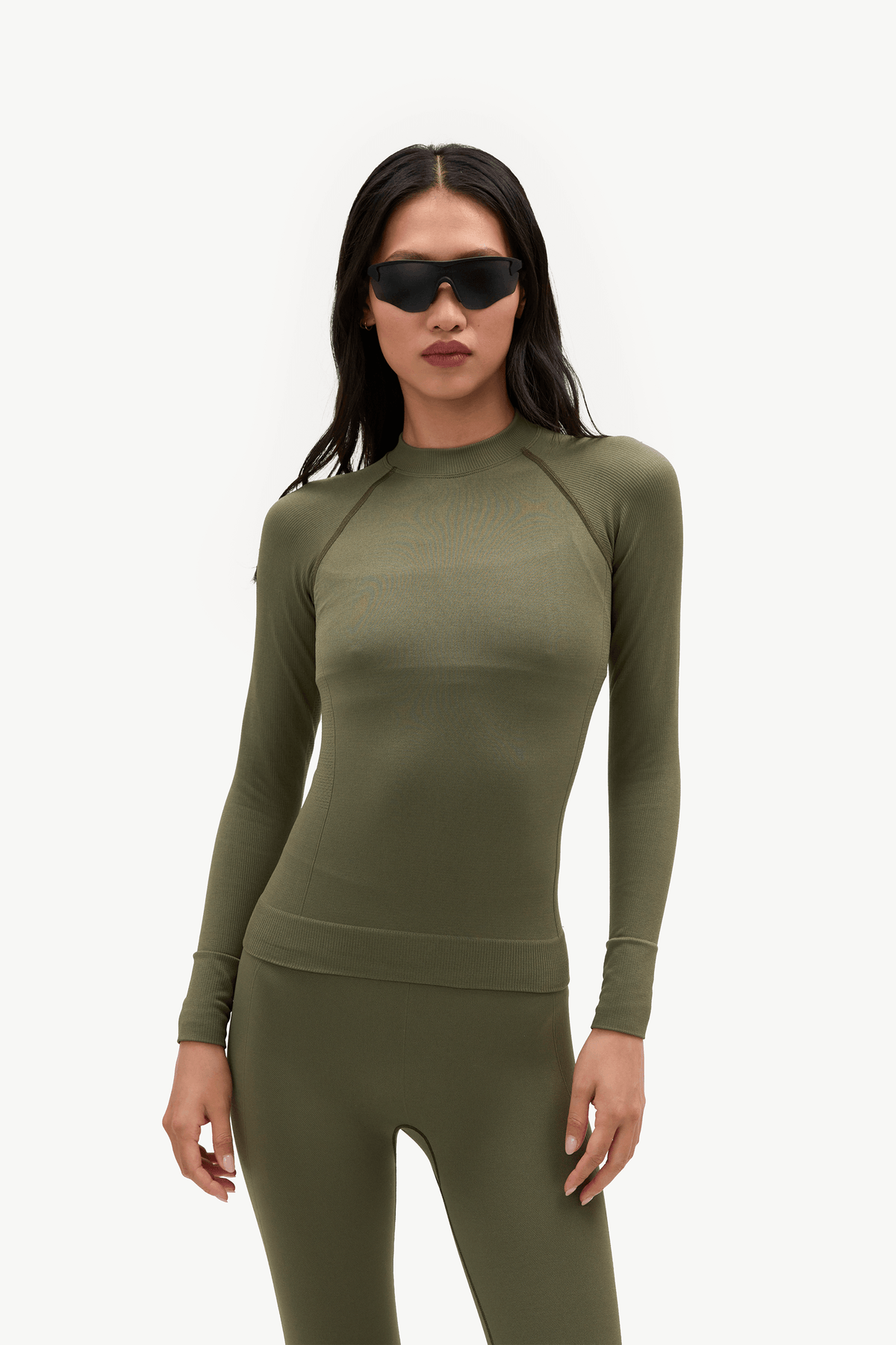 A woman modeling a matching olive green long-sleeve athletic top and leggings set against a plain light gray studio background.