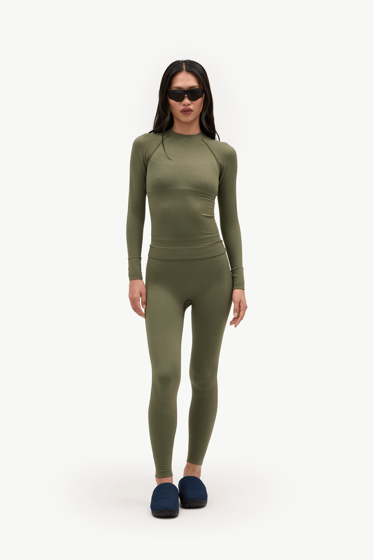 A model wearing a matching olive green long-sleeve top and high-waisted leggings set, styled with dark blue slippers and black sunglasses, against a plain white studio background.