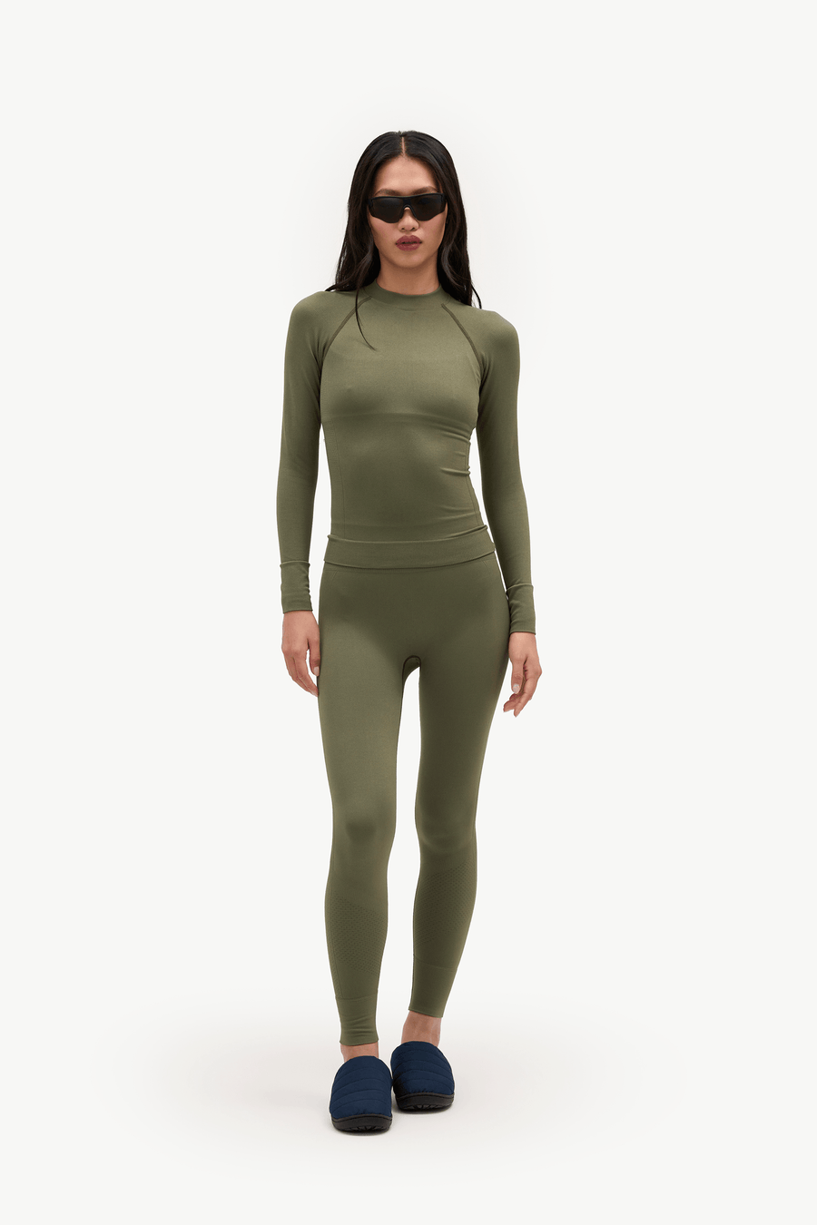 A model wearing a matching olive green long-sleeve top and high-waisted leggings set, styled with dark blue slippers and black sunglasses, against a plain white studio background.