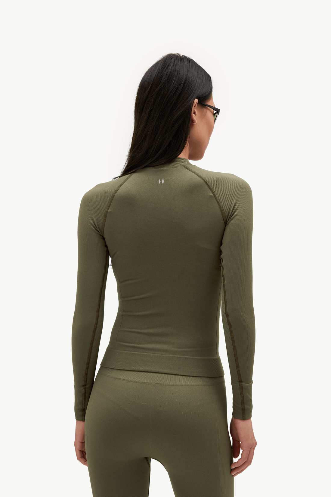 A woman viewed from the back wearing a fitted long-sleeve athletic top and matching leggings in an olive green color, against a plain light background.
