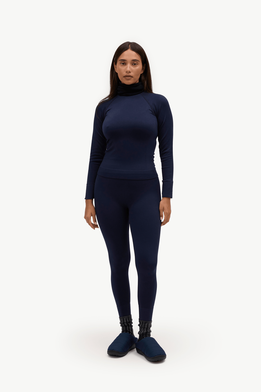 A woman modeling a matching navy blue activewear set consisting of a long-sleeve mock neck top and high-waisted leggings, paired with navy socks, against a plain white studio background.
