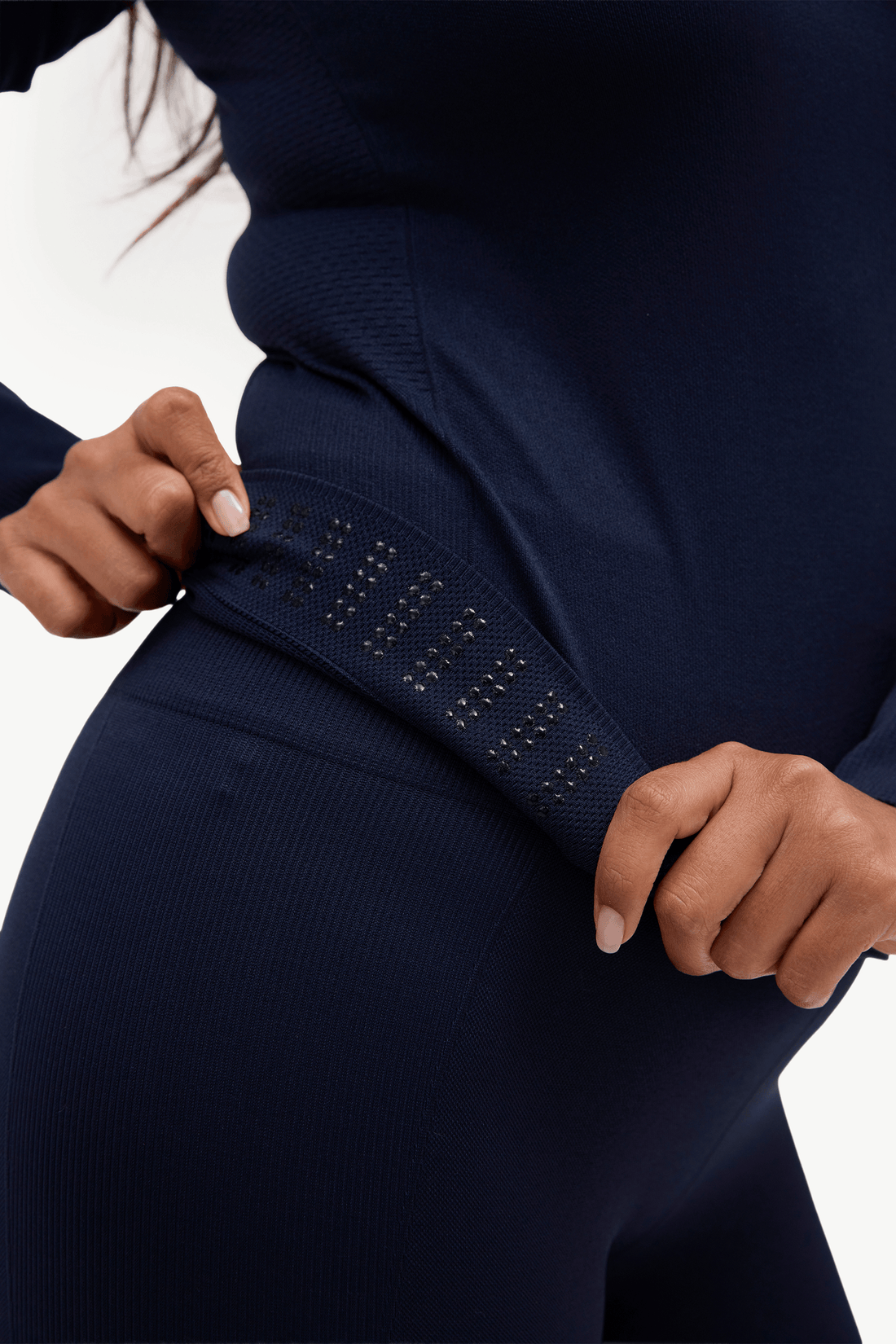 Close-up of a person wearing a dark navy ribbed knit jumpsuit or matching set, pulling the side seam slightly to show stretch and texture against a plain light background.