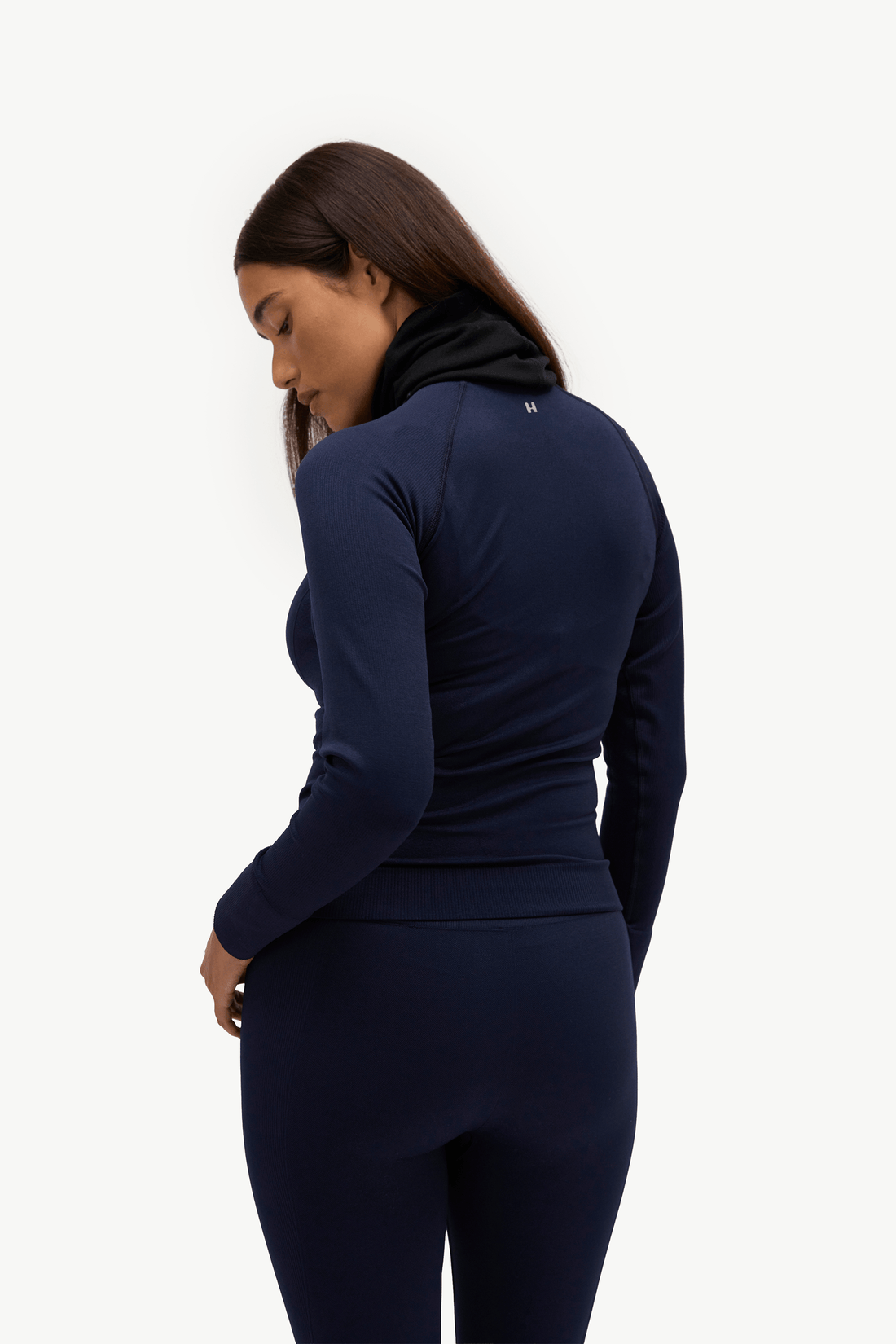 A woman wearing a navy blue long-sleeve athletic top and matching leggings, shown from a side-back angle against a plain white studio background.
