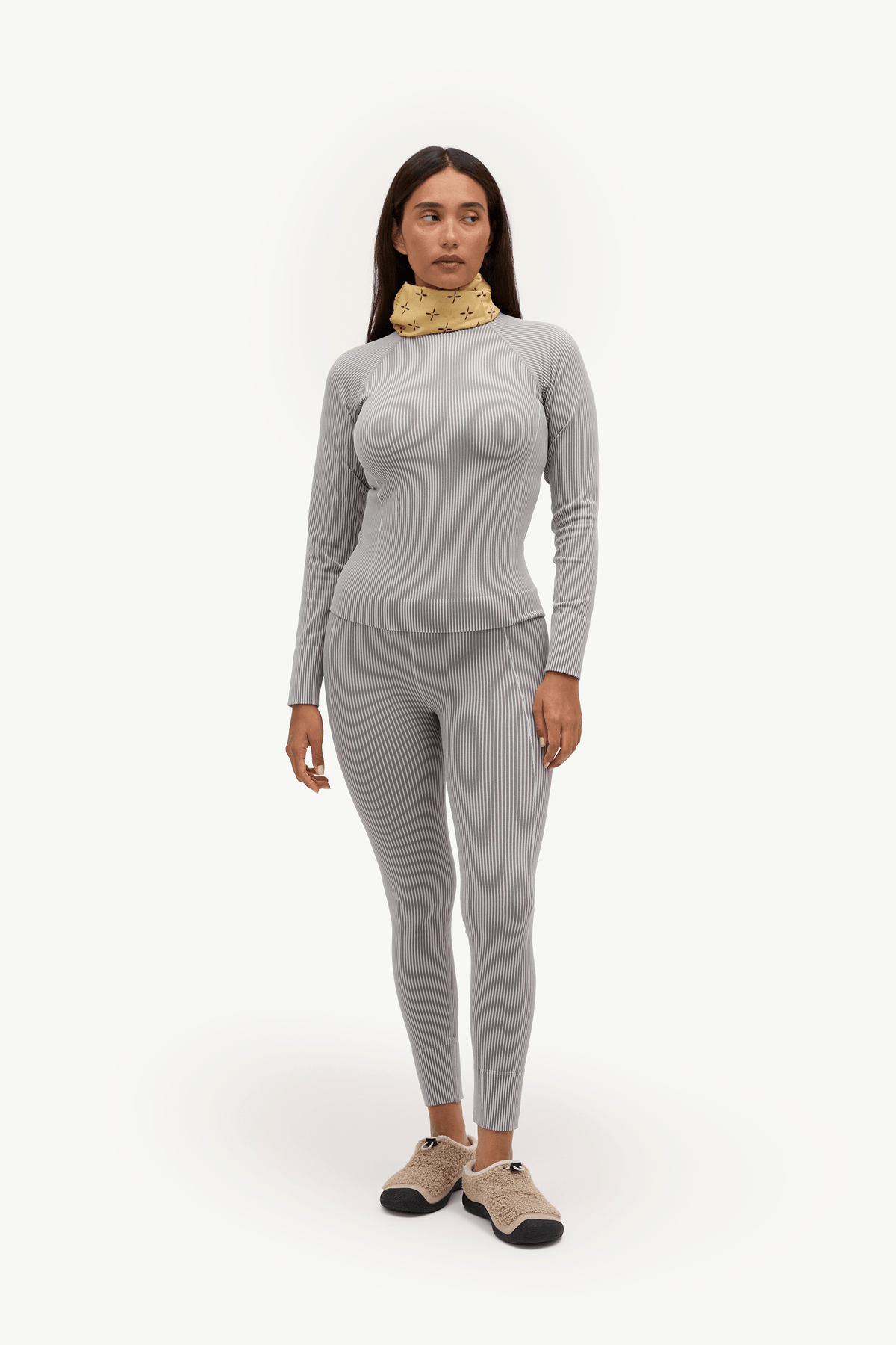 A woman modeling a light gray ribbed long-sleeve top and matching leggings set, styled with a textured gold neck warmer and beige slip-on shoes, against a plain white studio background.