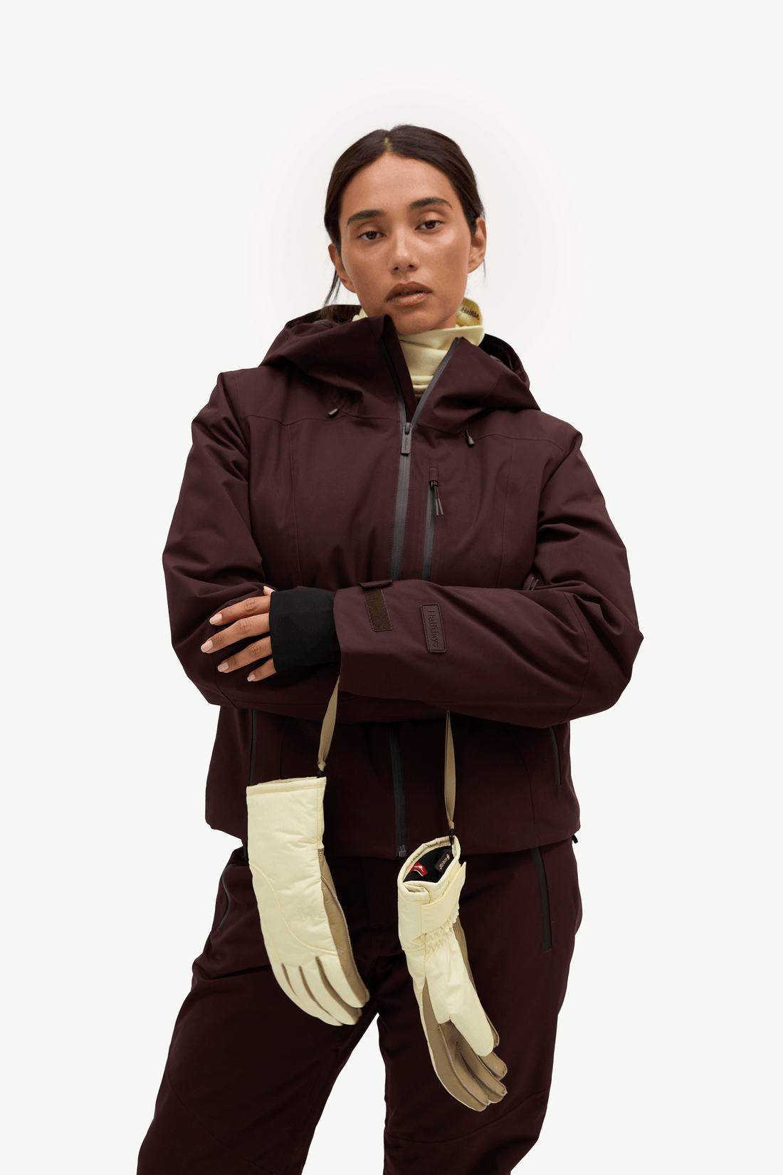 A model wearing a dark brown ski jacket and matching pants, accessorized with cream-colored gloves and a cream neck warmer, against a solid white studio background.