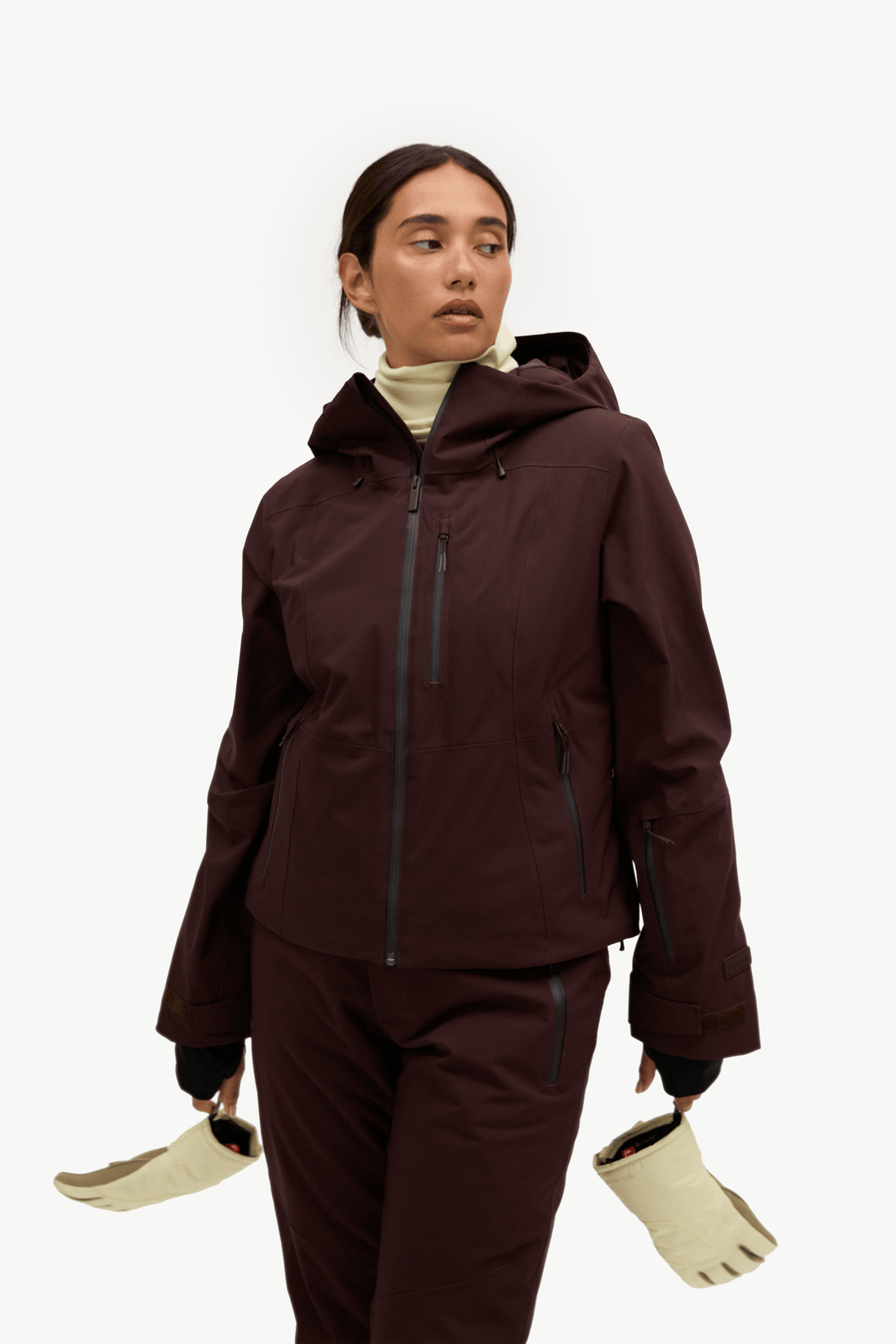 A person wearing a dark burgundy hooded ski jacket with black zippers and a black inner sleeve, holding gloves.