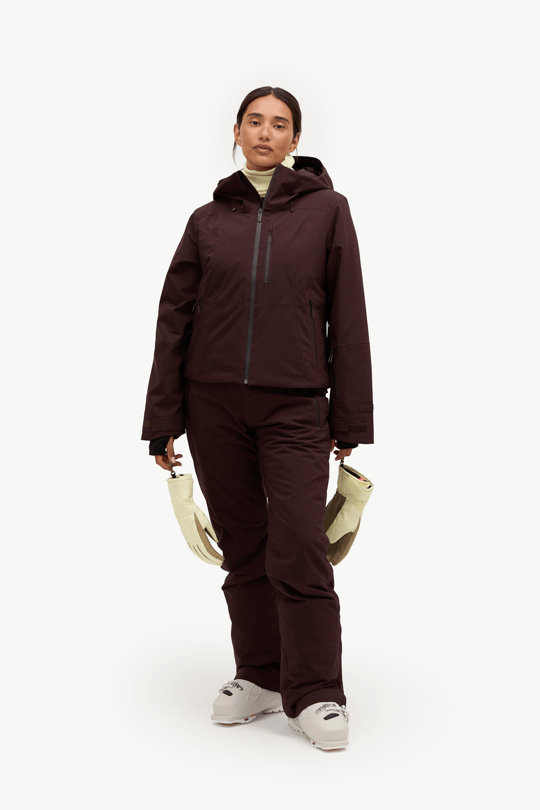 A woman modeling a dark brown ski jacket and matching ski pants, holding a pair of cream-colored gloves against a plain white studio background.
