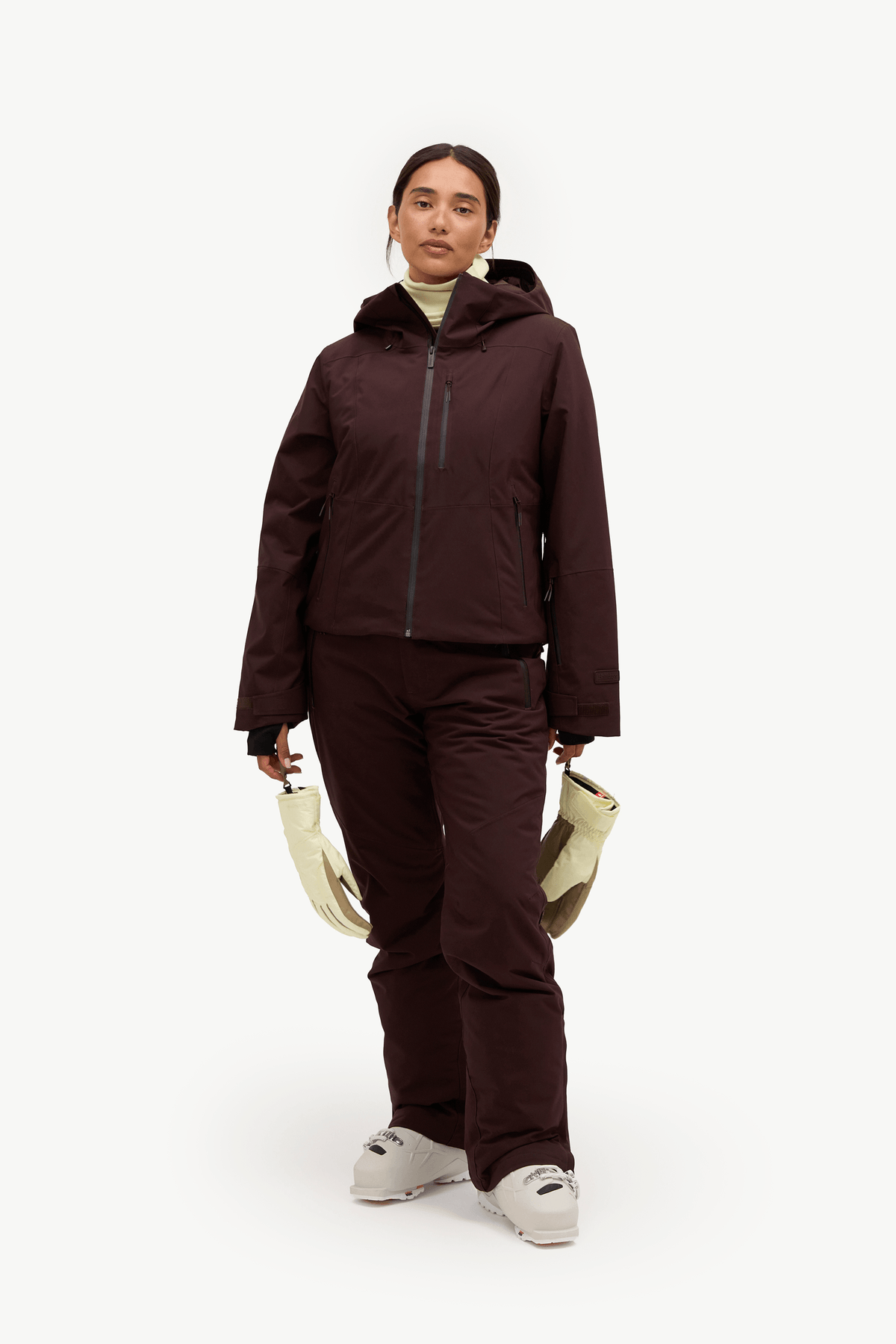 A woman modeling a dark brown ski jacket and matching ski pants, holding a pair of cream-colored gloves against a plain white studio background.