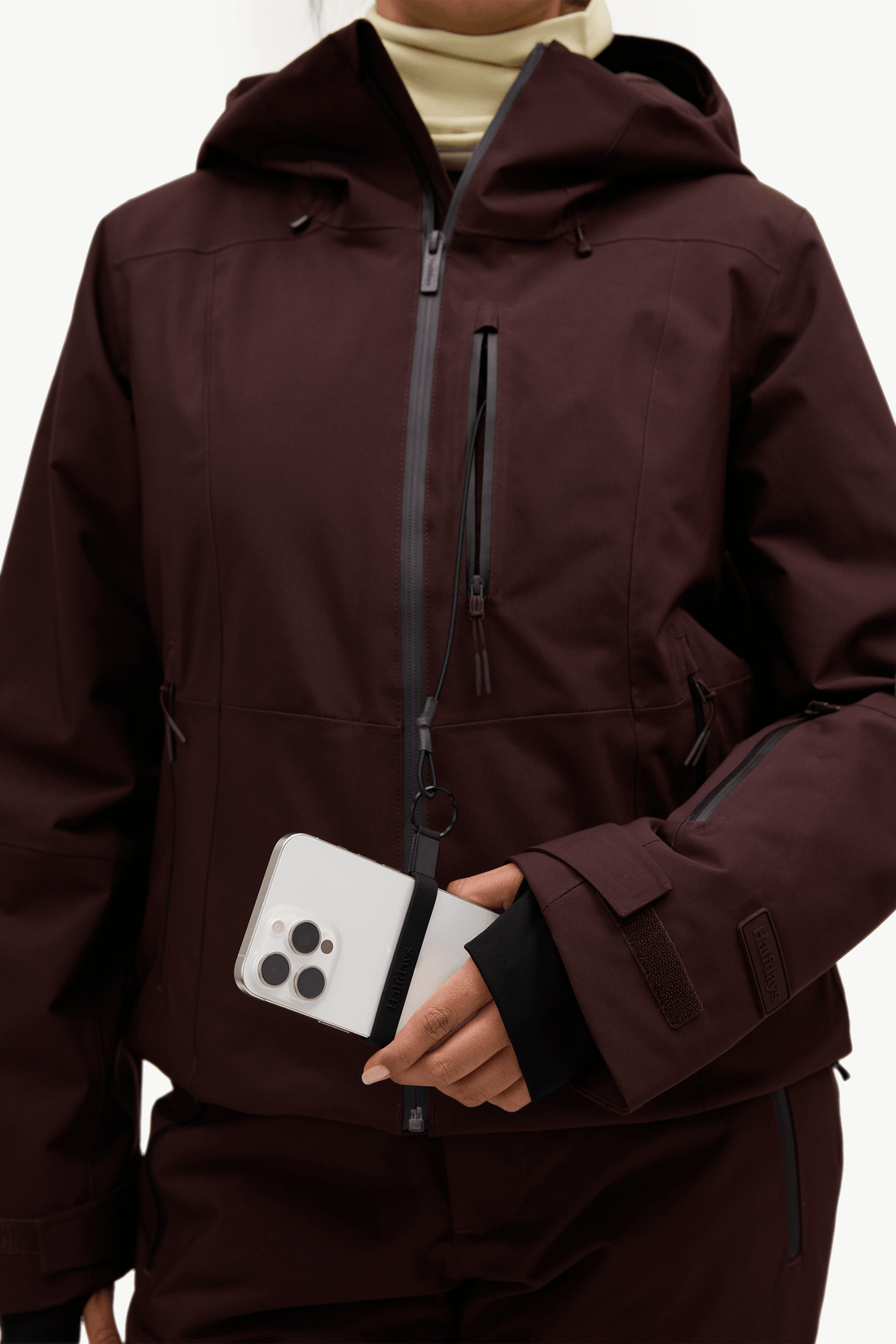 A close-up of a person wearing a dark burgundy hooded ski jacket with black zippers and a black inner sleeve, holding a white smartphone in front of the jacket pocket.