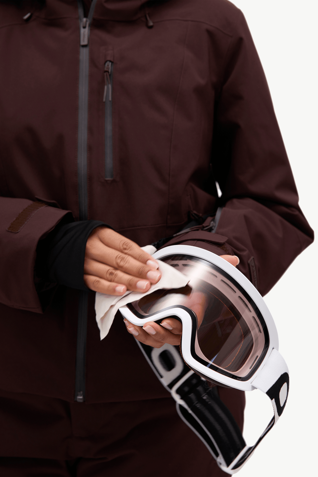 A close-up of a person wearing a dark burgundy hooded ski jacket with black zippers and a black inner sleeve, holding goggles.