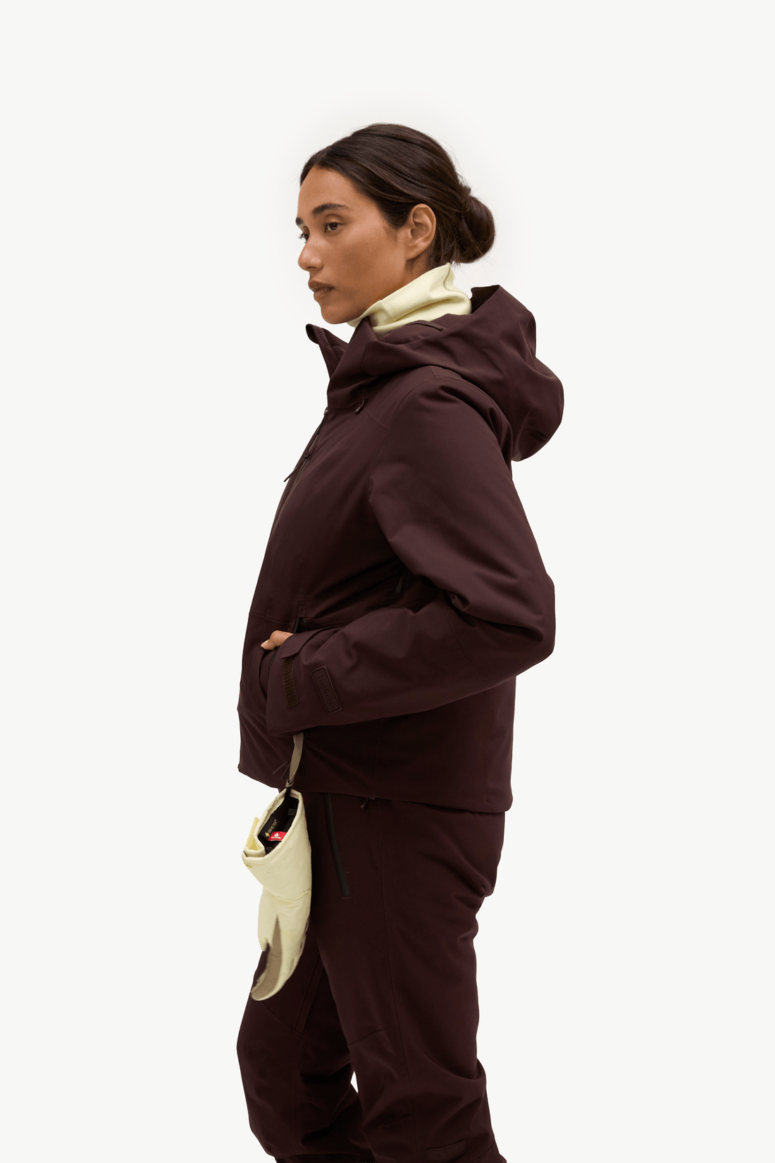 A woman in profile wearing a dark plum-brown hooded jacket with a cream inner collar and matching dark pants, standing against a plain light gray studio background.