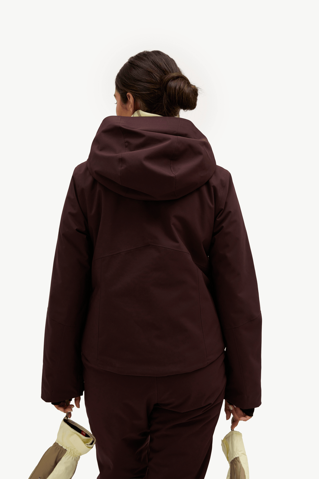 A woman viewed from behind wearing a dark plum-brown hooded jacket and matching pants, holding a light beige bag against a plain white studio background.