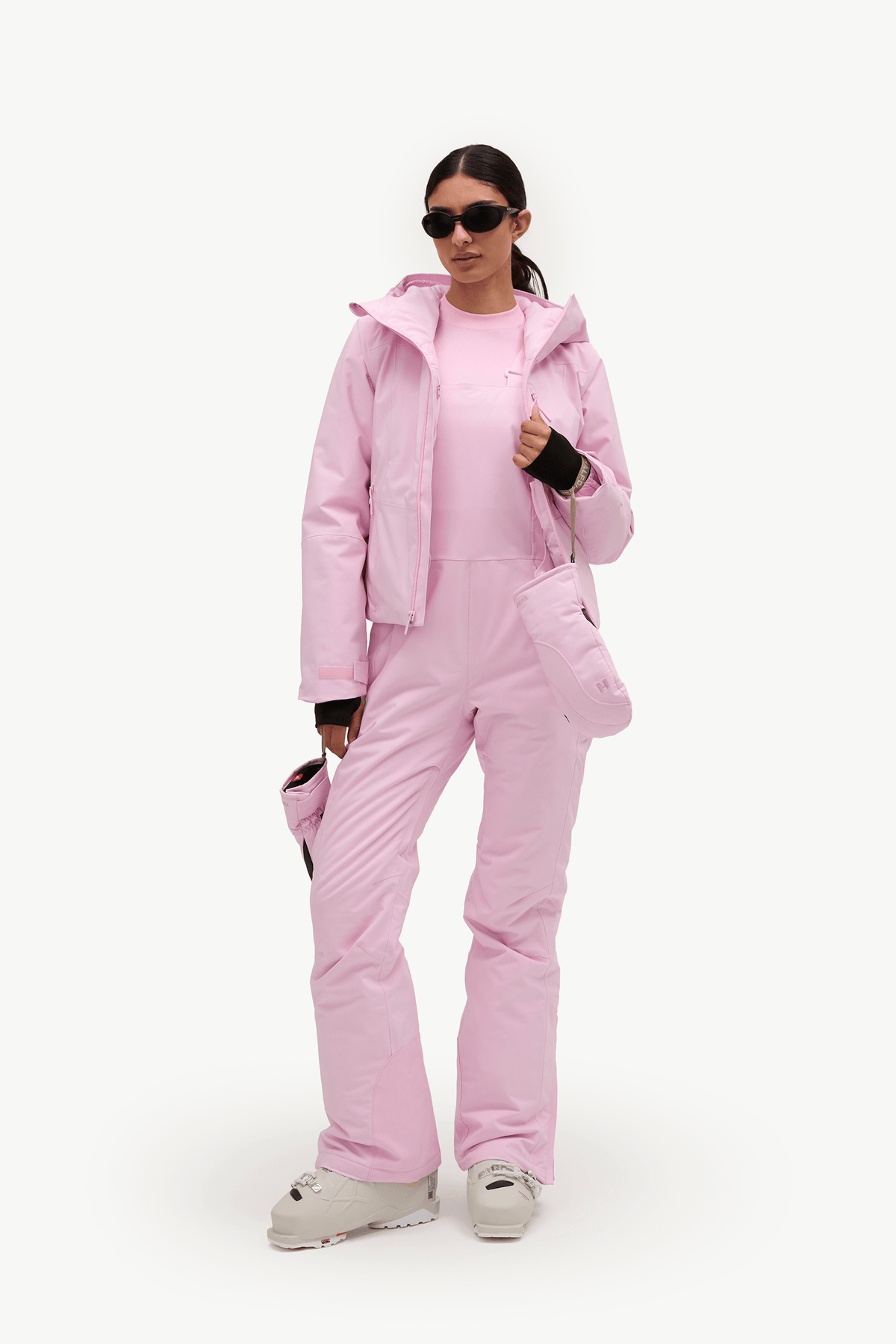 A model wearing a light pink ski outfit consisting of a hooded jacket, a matching top, and ski pants, paired with white sneakers and black sunglasses, set against a plain white studio background.