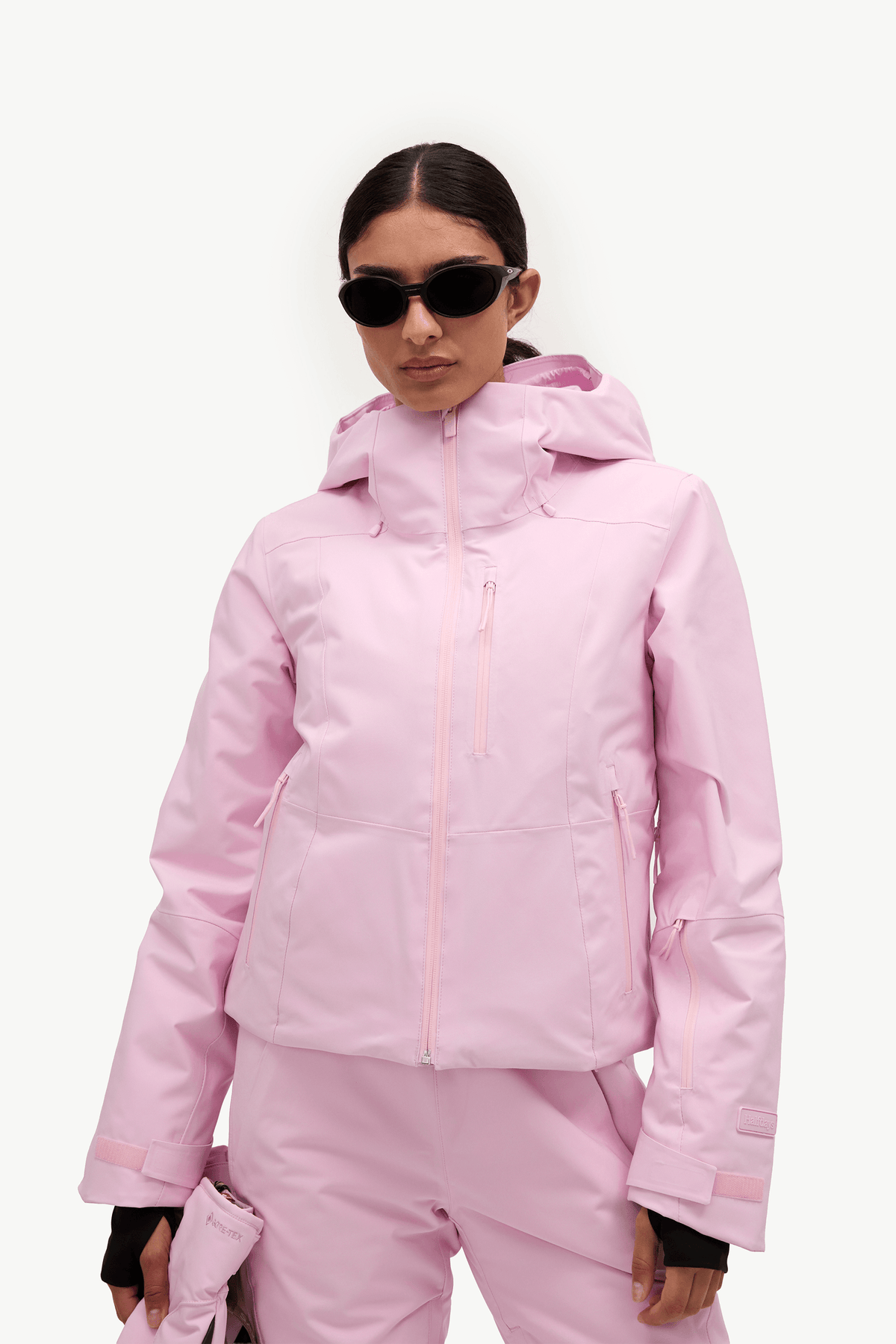 A model wearing a light pink ski jacket and matching ski pants, accessorized with black sunglasses, against a clean white studio background.