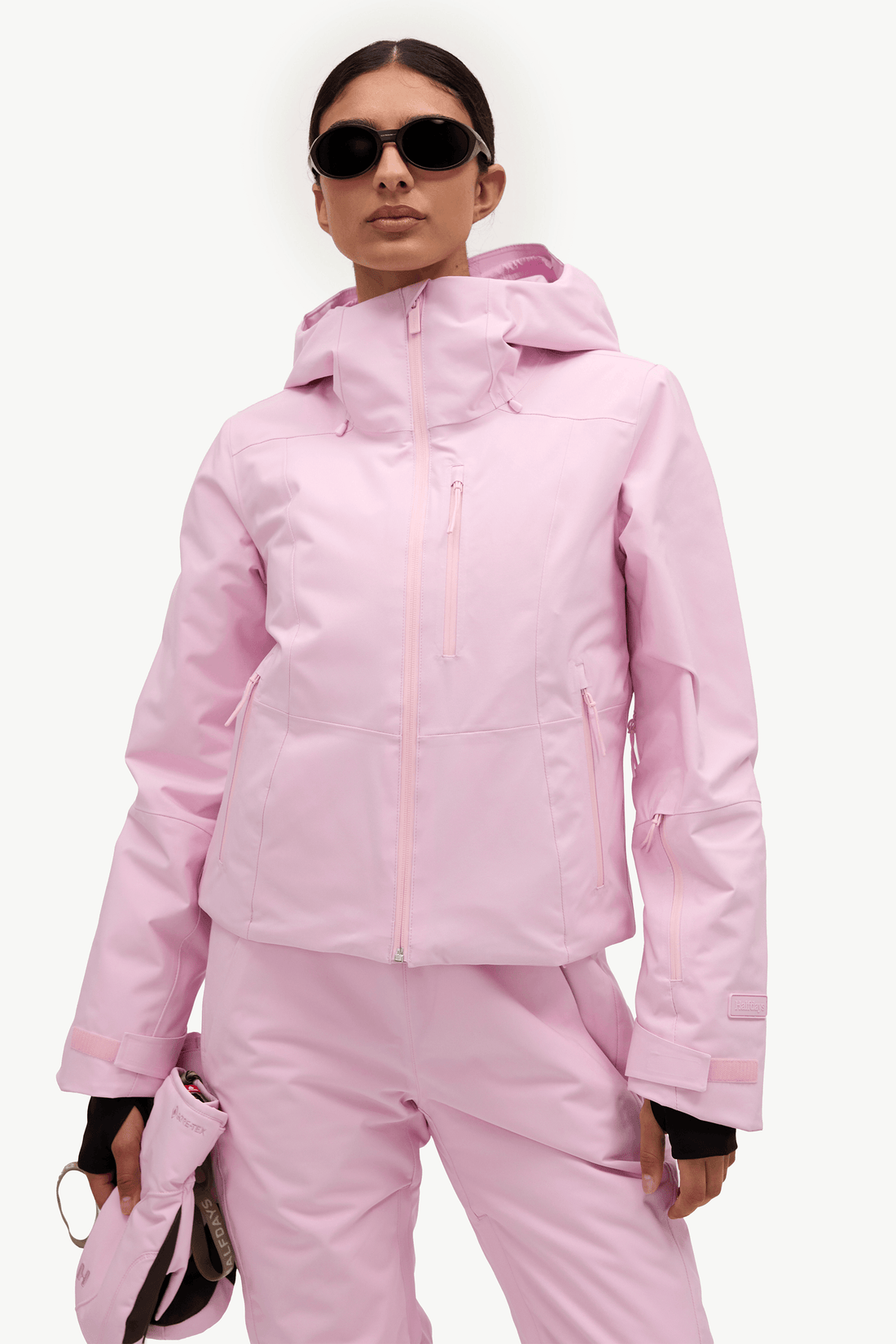 A model wearing a light pink ski jacket and matching ski pants, holding a matching pink ski boot in her hand against a plain white studio background.