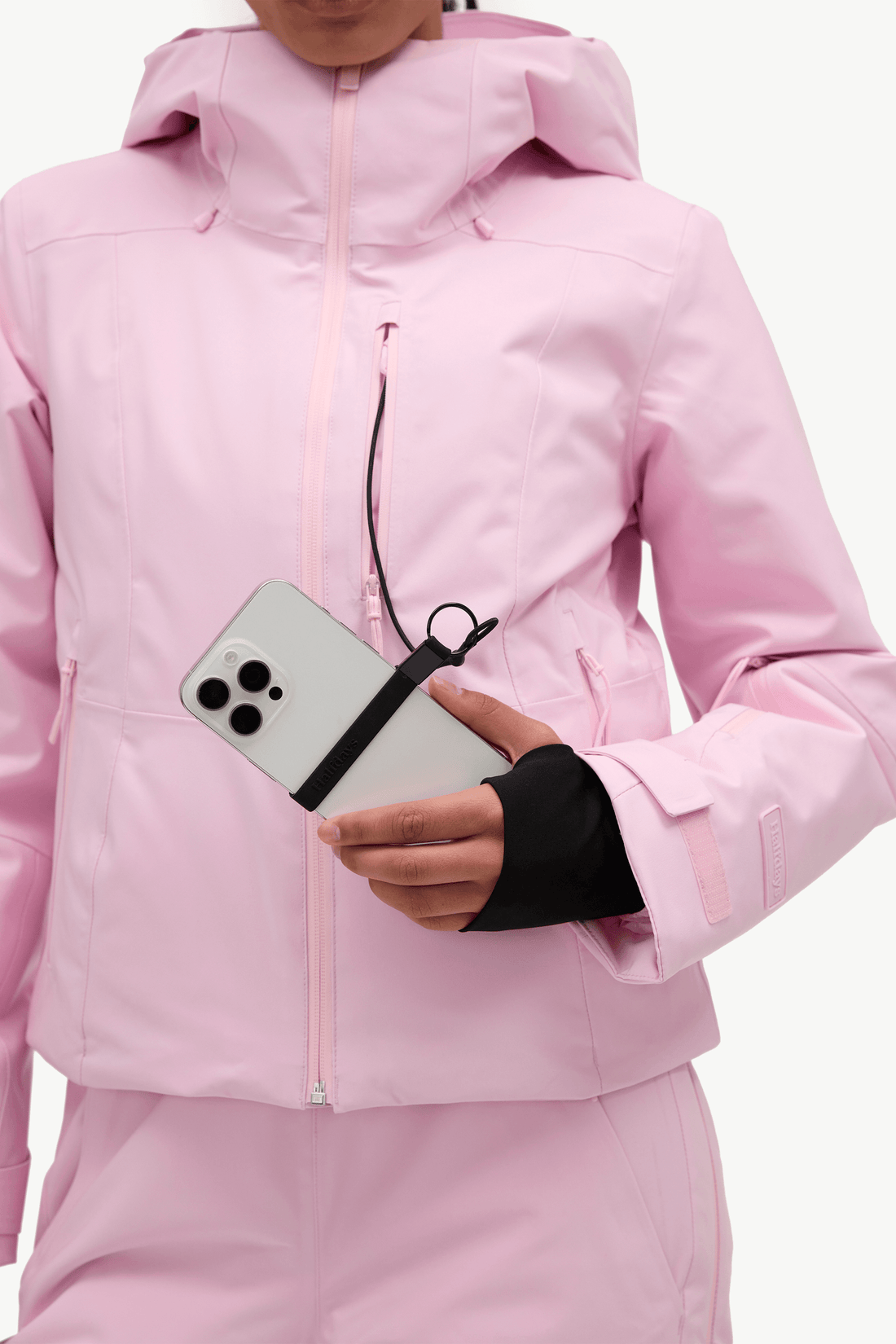A close-up of a person wearing a light pink ski jacket and matching pink pants, holding a silver smartphone with a black wrist strap in one hand and wearing a black wrist sleeve in the other.