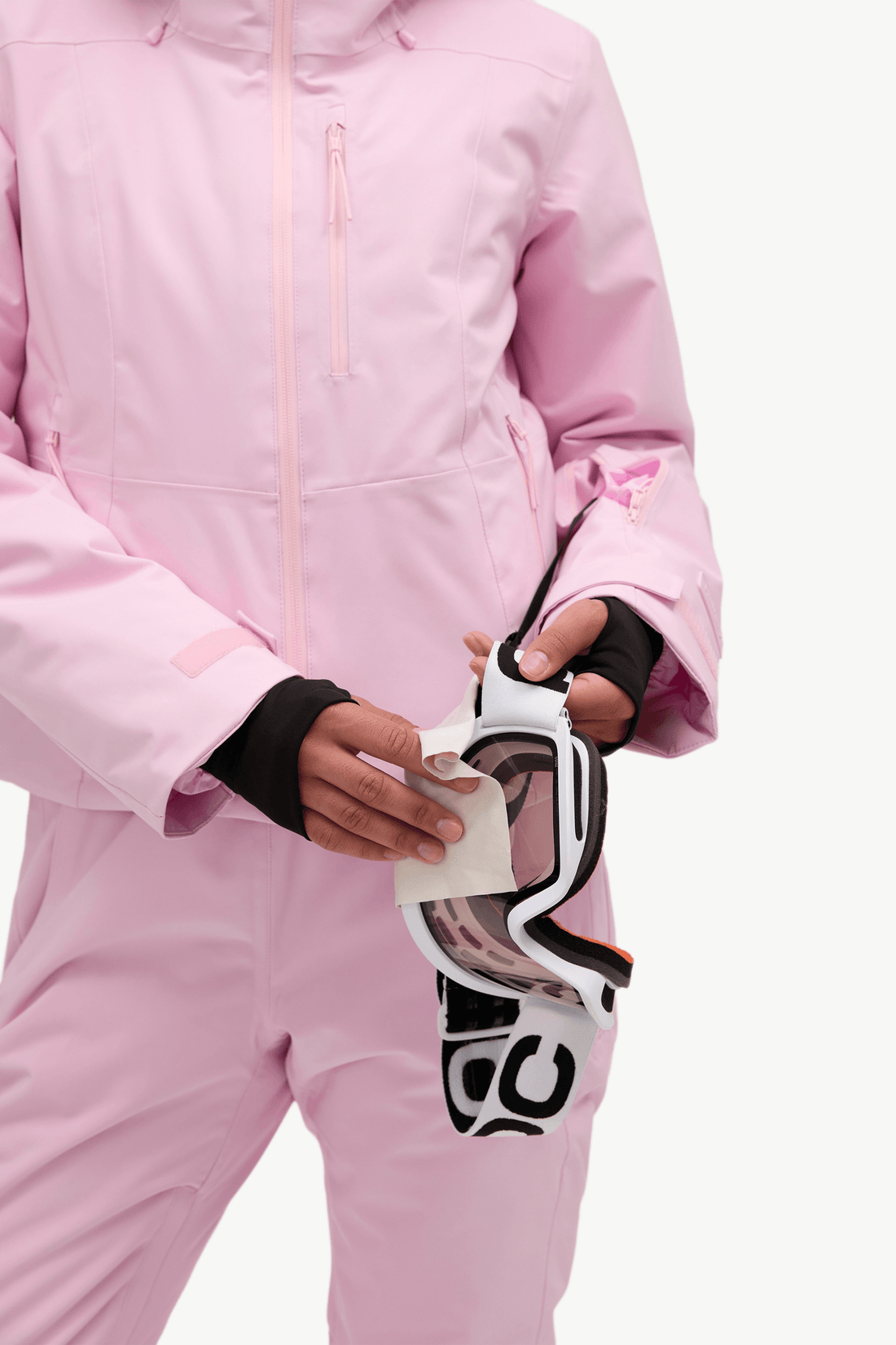 A person wearing a light pink ski jacket and matching pants holds a pair of white, black, and pink ski goggles against a clean white background.