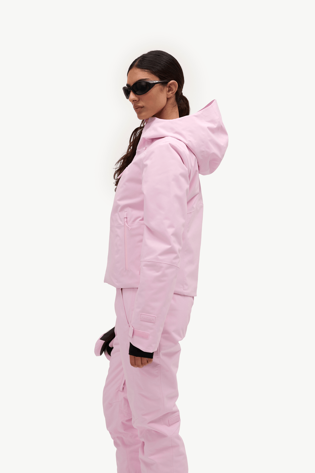 A woman wearing a light pink ski jacket and matching ski pants, shown in profile against a plain white studio background.