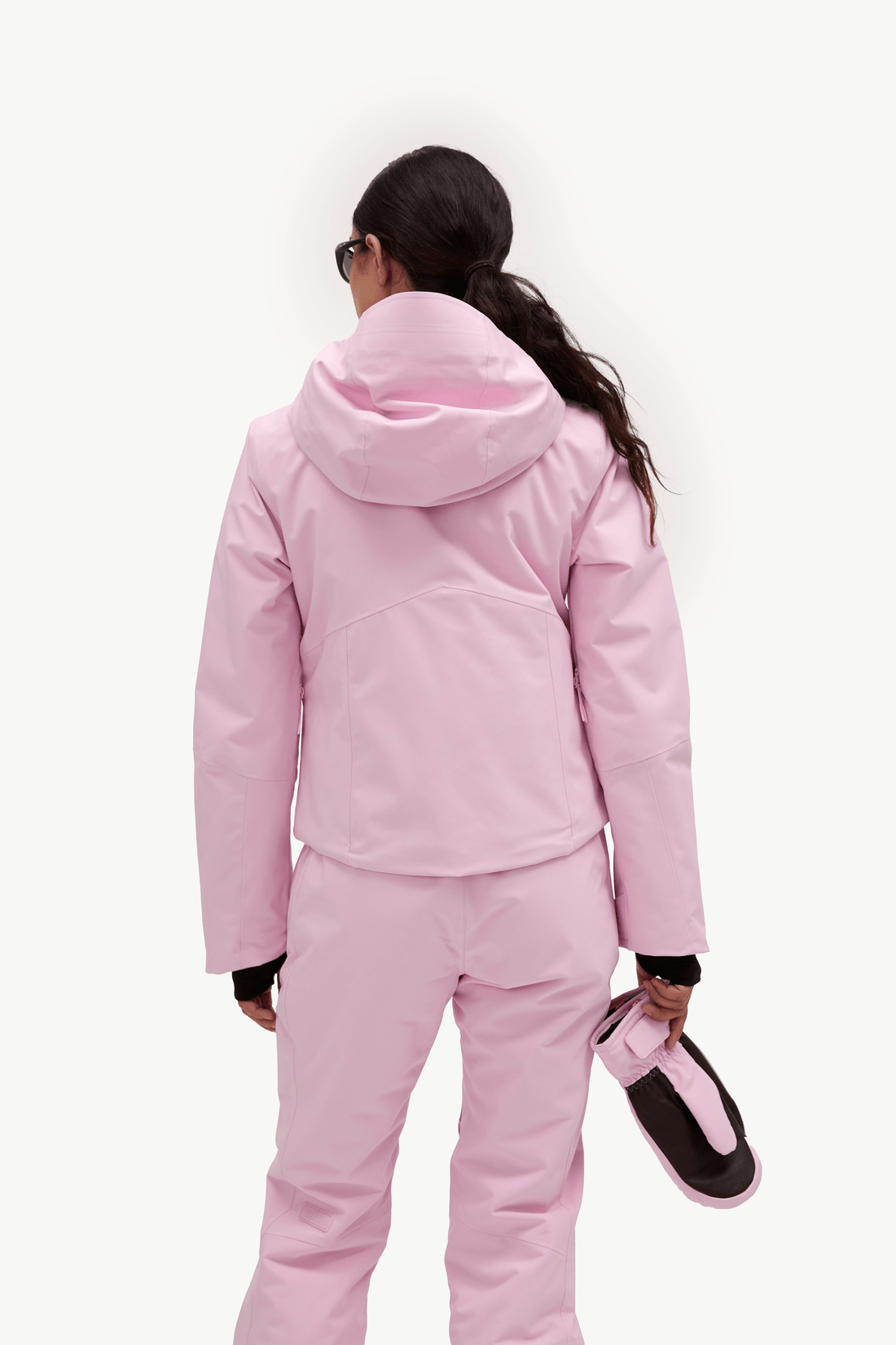 A model from behind wearing a light pink ski jacket and matching pants, holding a pink and black ski boot against a plain white background.