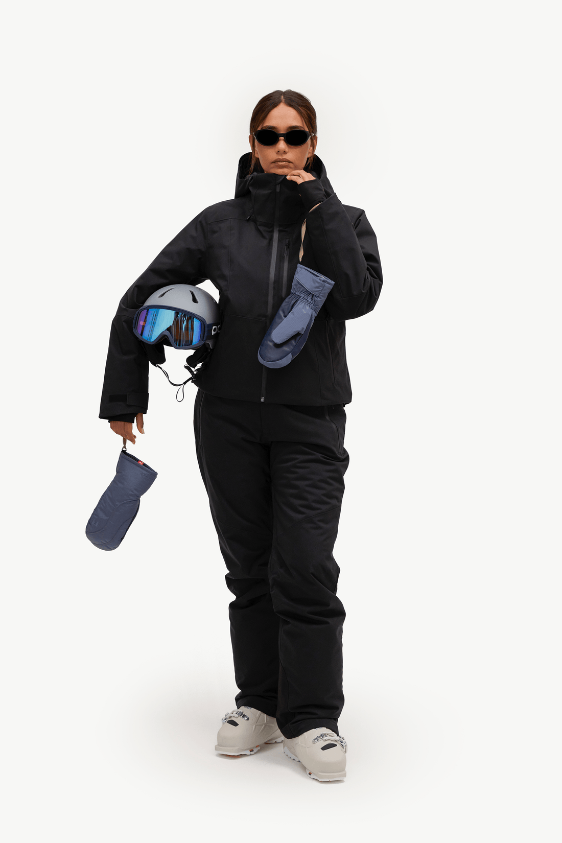 A woman modeling a black ski jacket and matching black ski pants, holding a ski helmet with blue-tinted goggles, a dark blue glove, and light gray sneakers against a plain white studio background.