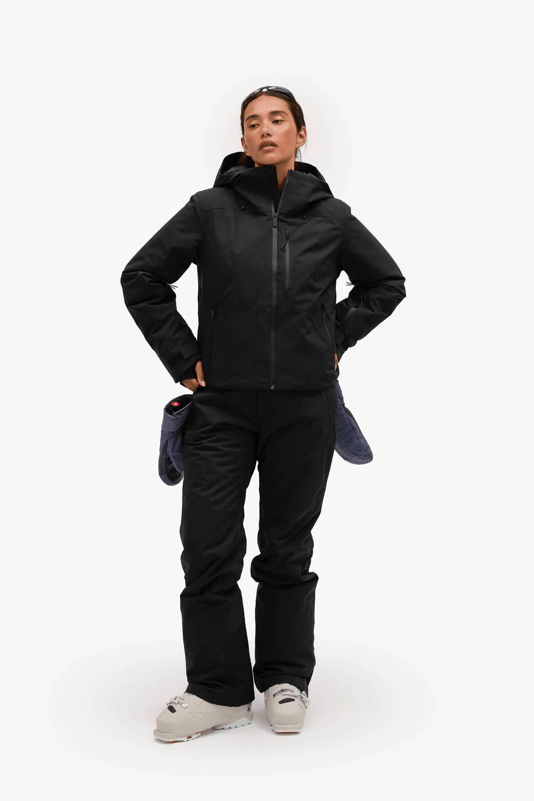 A woman wearing a black hooded jacket and matching black pants, shown in profile against a plain light background. Sunglasses are resting on her head.