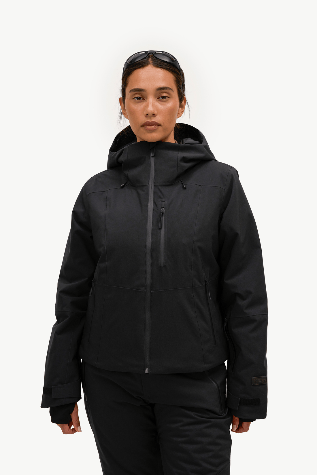 A woman modeling a black hooded ski jacket with a full front zipper and zippered chest pocket, paired with black ski pants, against a plain white studio background.