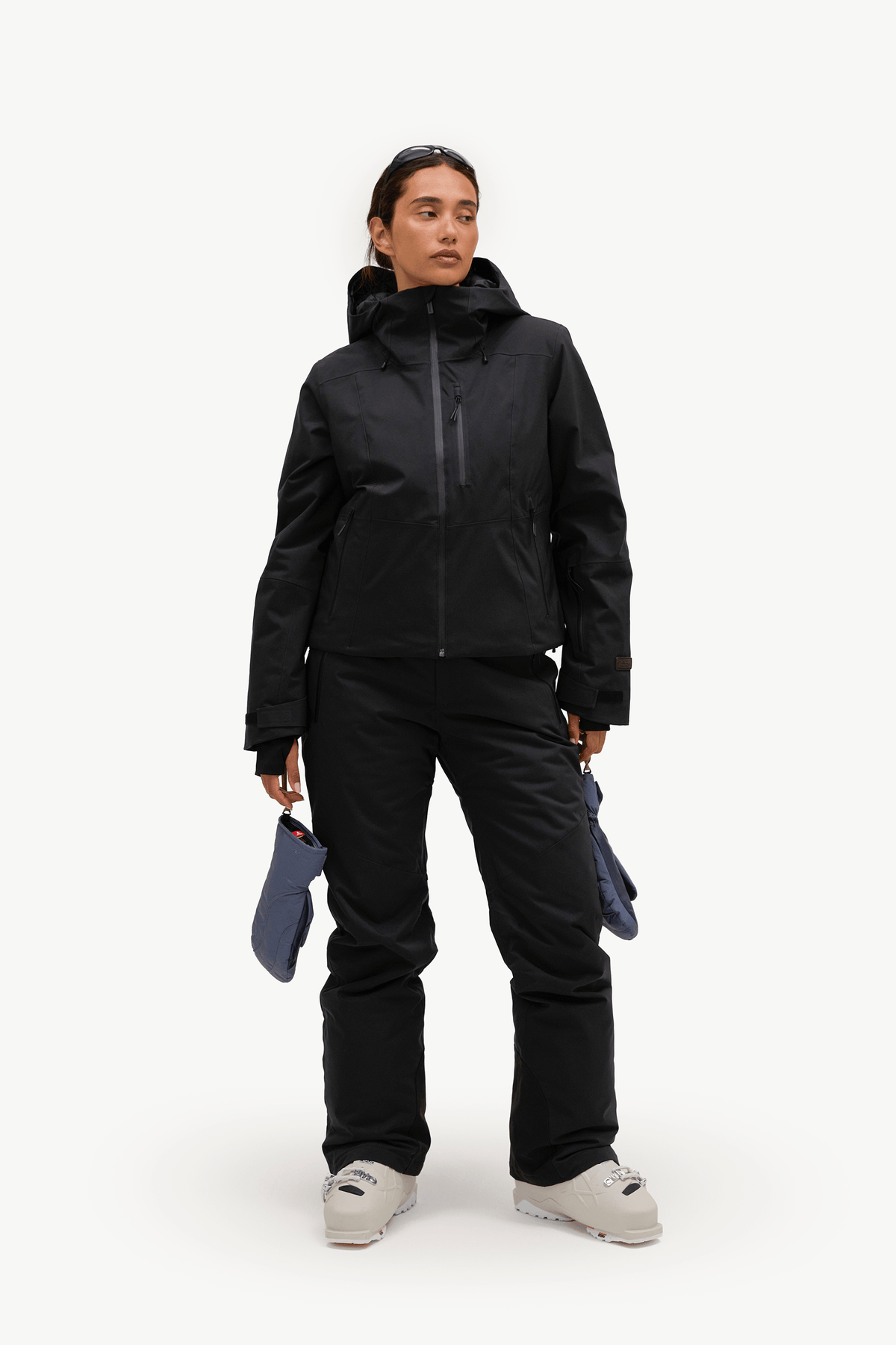 A model stands against a plain white studio background wearing a black ski jacket and black ski pants, holding a pair of blue gloves.