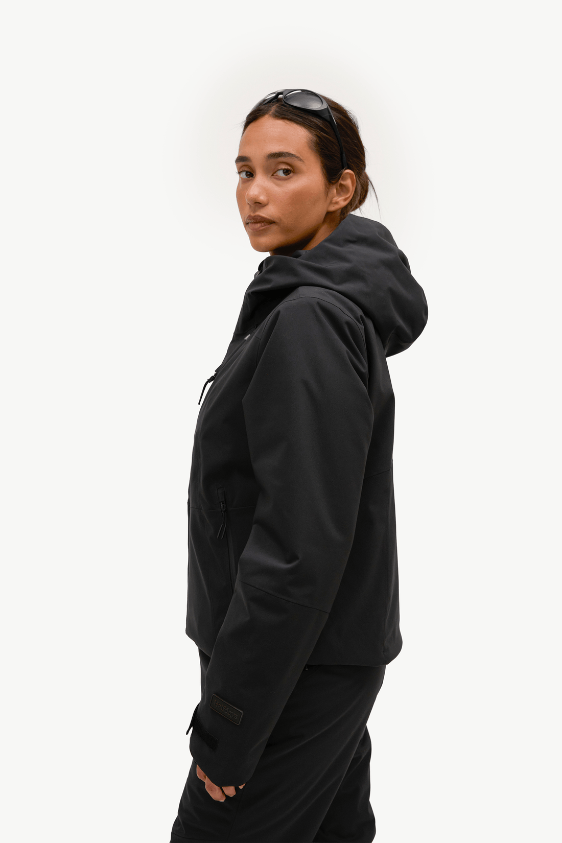 A woman wearing a black hooded jacket and matching black pants, shown in profile against a plain light background. Sunglasses are resting on her head.