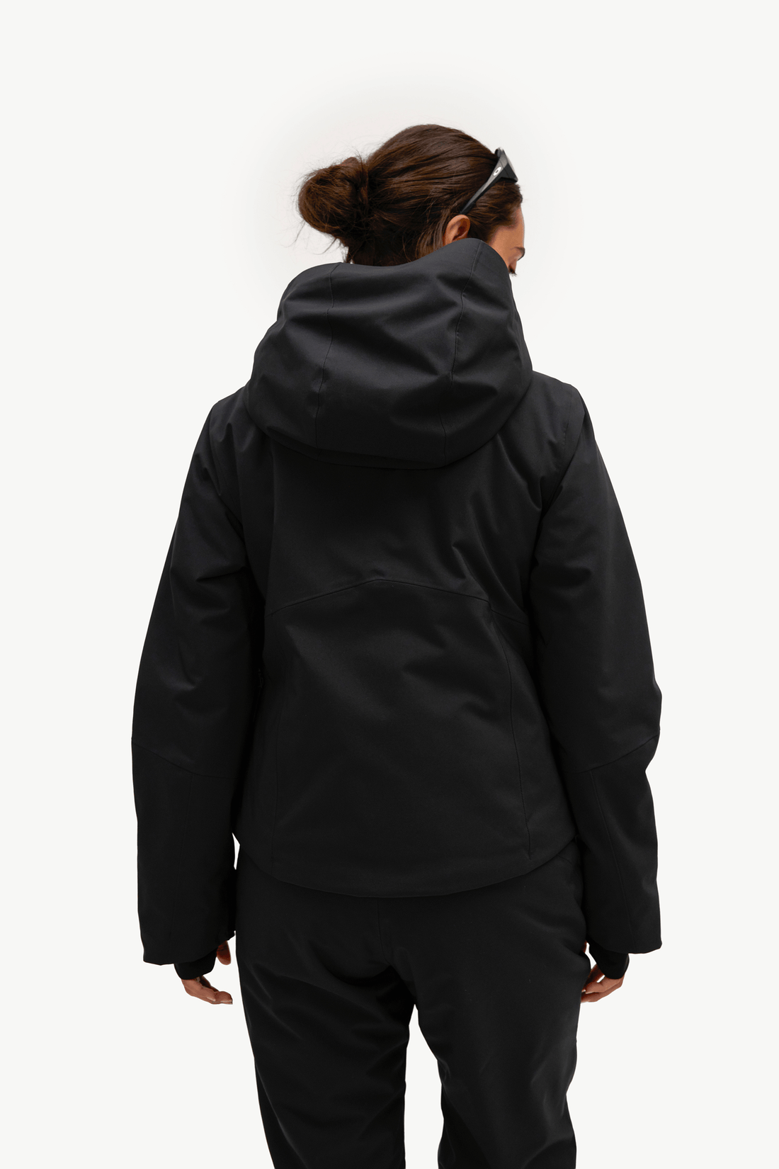 A woman viewed from behind wearing a black hooded jacket and matching black pants against a plain white studio background.
