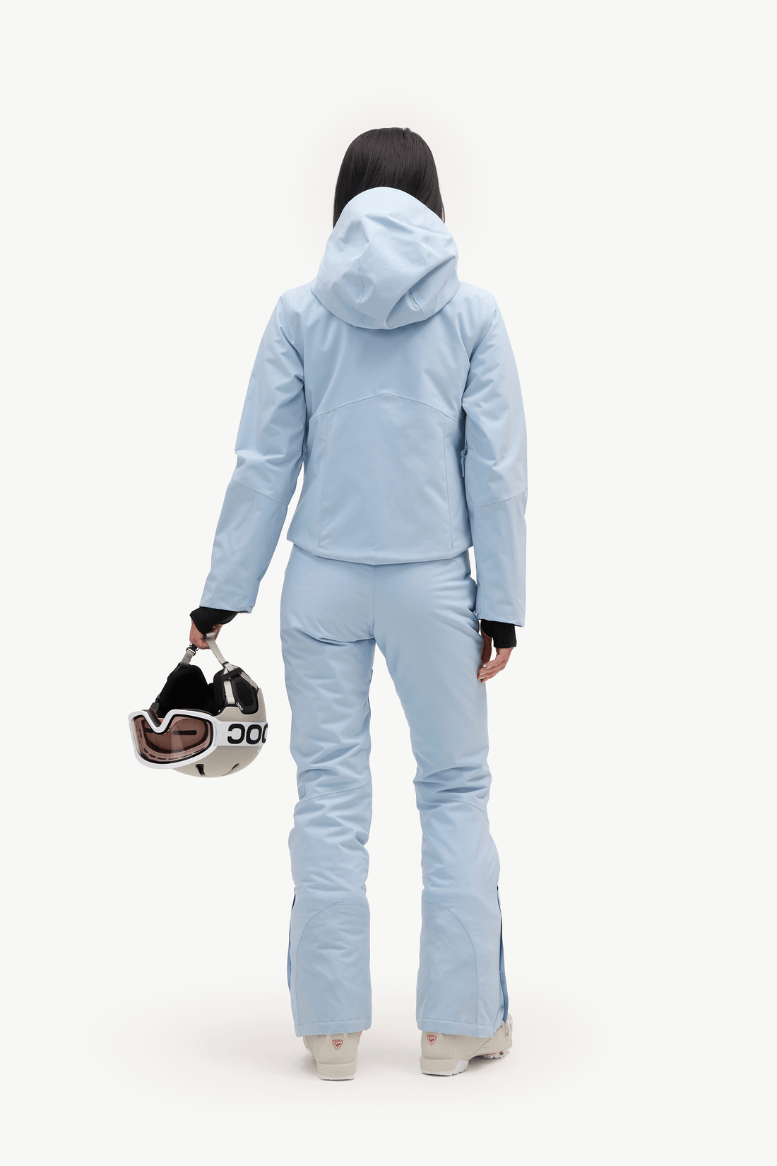 A model from behind wearing a light blue ski jacket and matching pants, holding a black and white ski helmet with goggles, against a solid white background.