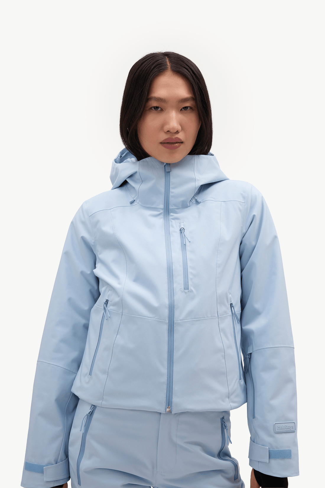 A model wearing a light blue hooded ski jacket and matching light blue ski pants against a clean white studio background.
