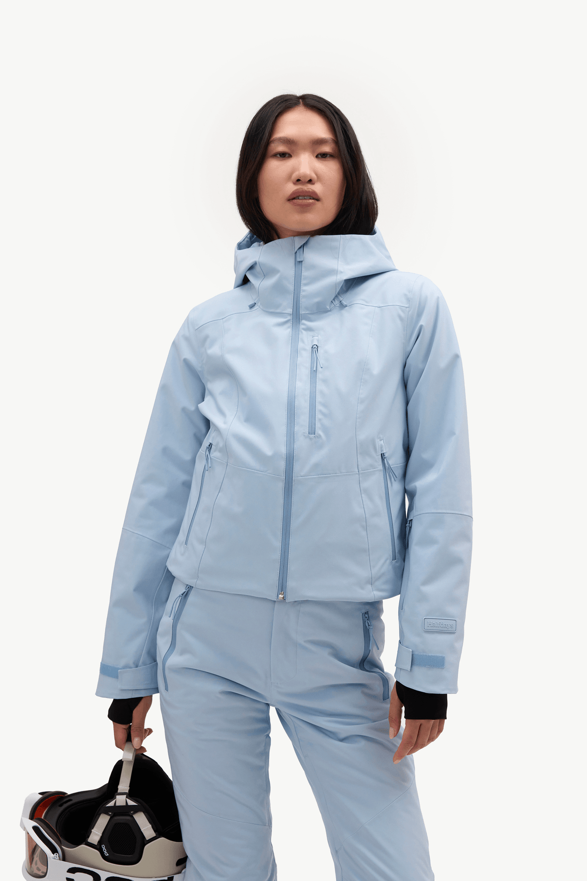 A model wearing a light blue ski jacket and matching pants, holding a ski helmet against a plain white studio background.