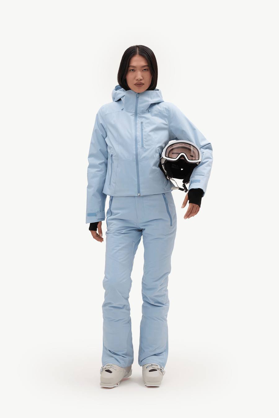 A model wearing a light blue ski jacket and matching pants, holding a ski helmet against a plain white studio background.