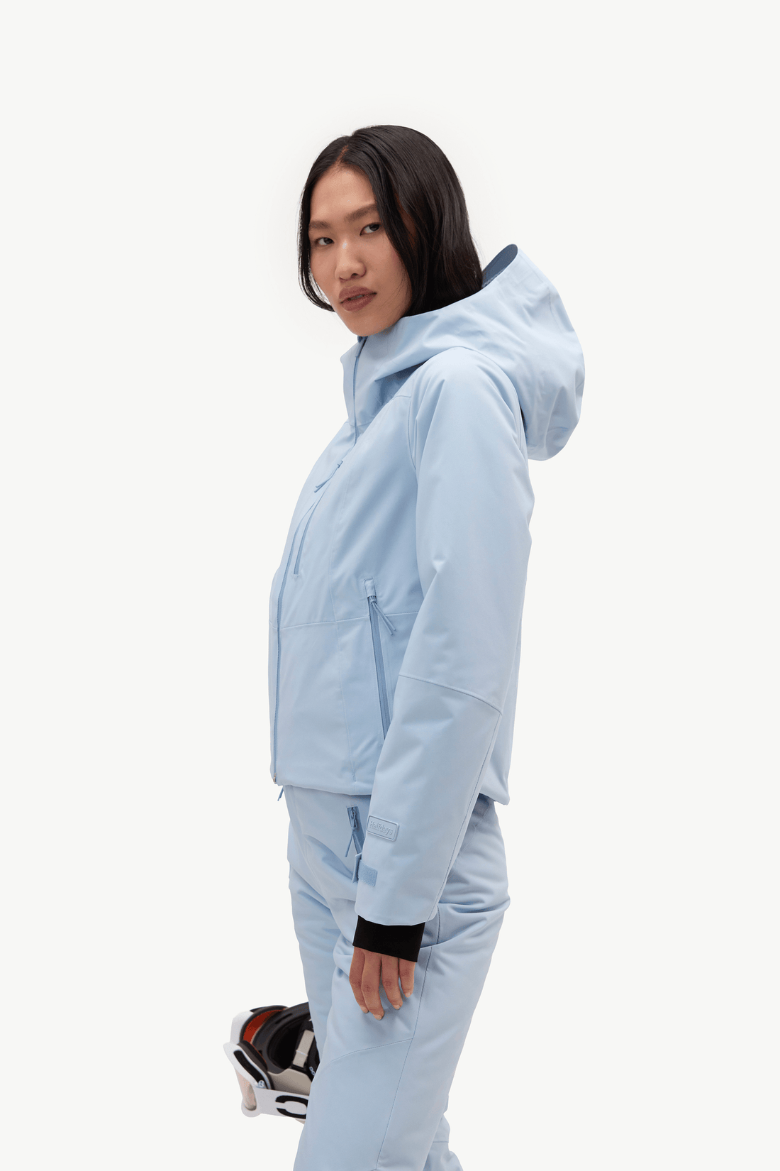 A model wearing a light blue hooded jacket and matching light blue pants, standing against a plain white studio background and holding a pair of ski goggles.