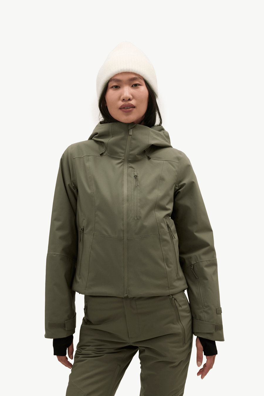 A model stands against a plain white studio background wearing an olive green ski jacket and matching ski pants, with a cream knit beanie and light-colored sneakers.