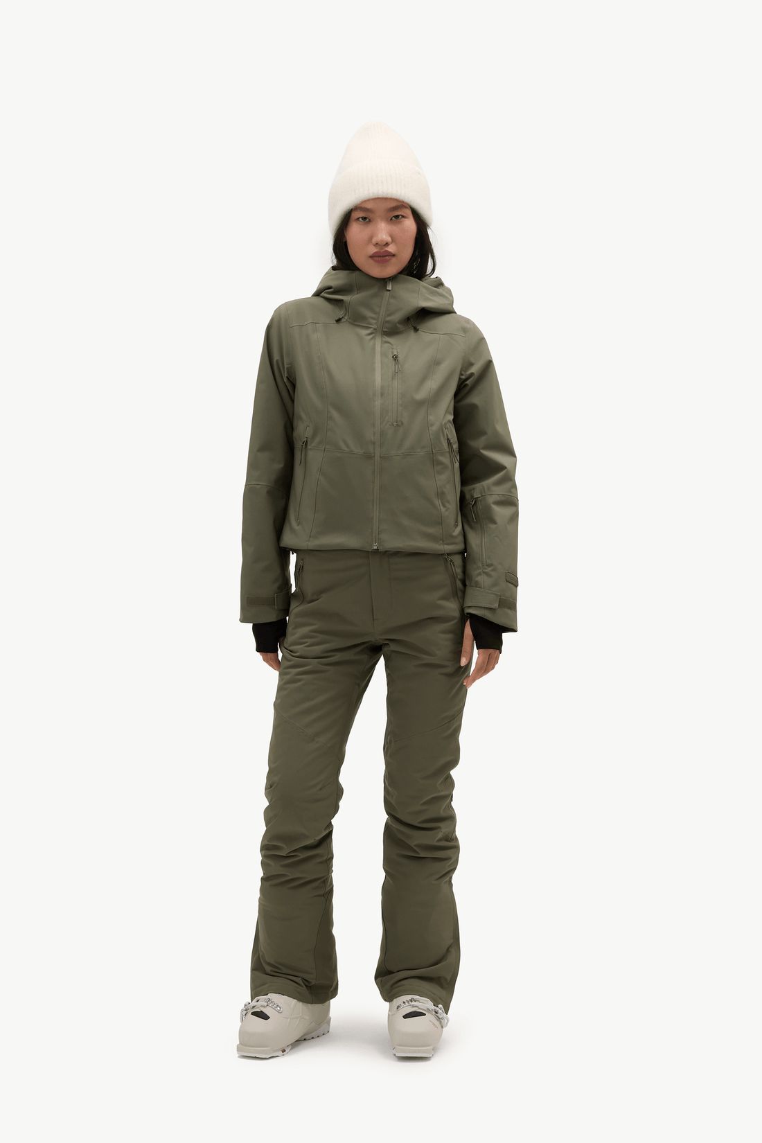 A model stands against a plain white studio background wearing an olive green ski jacket and matching ski pants, with a cream knit beanie and light-colored sneakers.