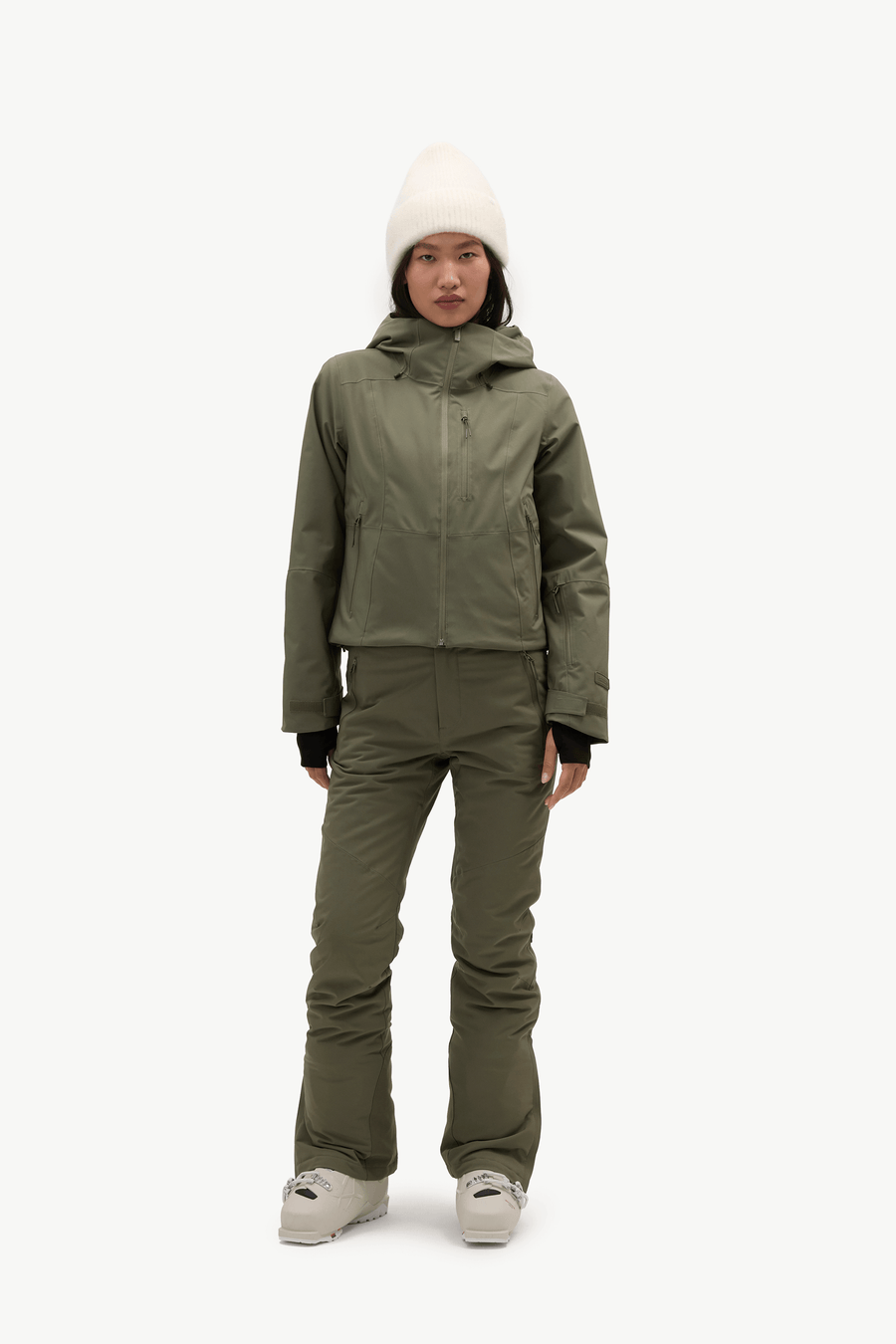 A model stands against a plain white studio background wearing an olive green ski jacket and matching ski pants, with a cream knit beanie and light-colored sneakers.