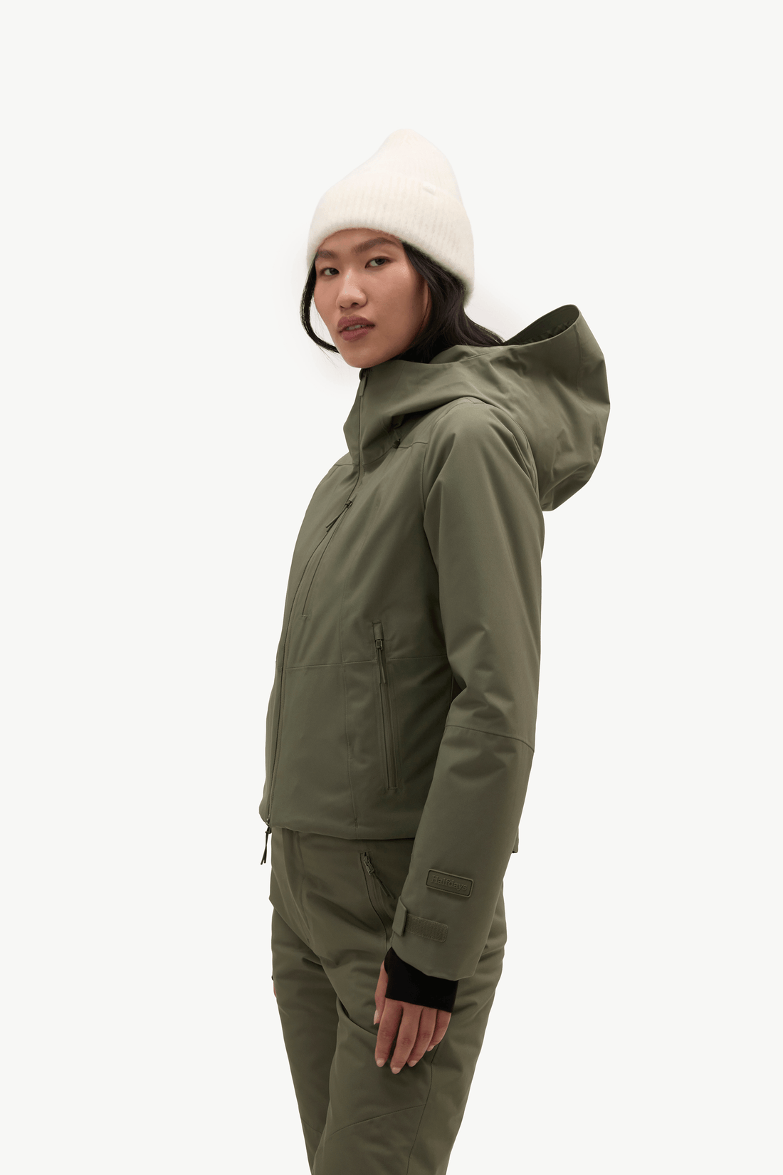 A model stands against a plain white studio background wearing an olive green ski jacket and matching ski pants, with a cream knit beanie and light-colored sneakers.