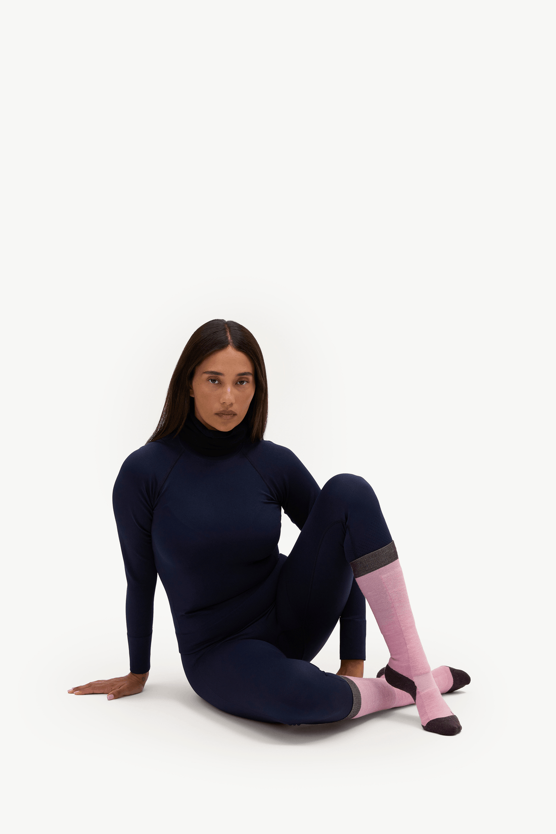 A woman sits on a white studio floor wearing a dark navy long-sleeve athletic turtleneck and matching leggings, paired with light pink knee-high socks.