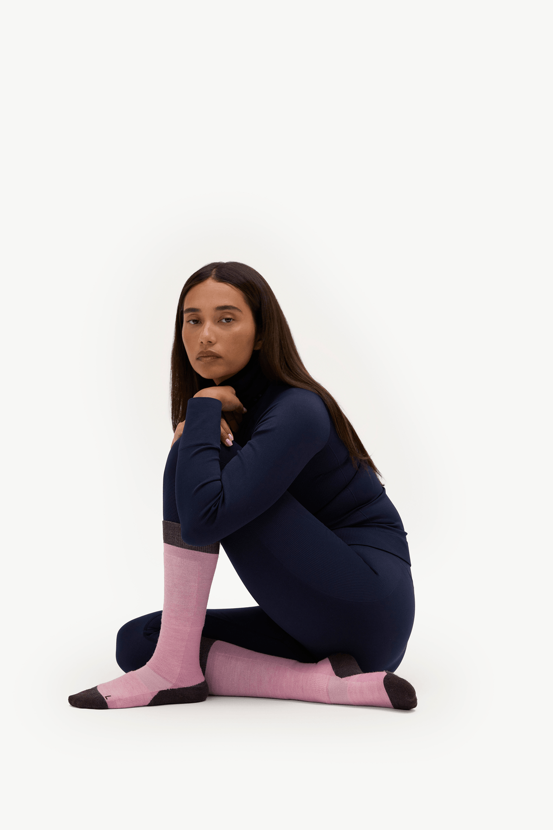 A woman sits cross-legged on a clean white studio background, wearing a dark navy long-sleeve top, matching navy leggings, and light pink socks with black toe and heel caps.