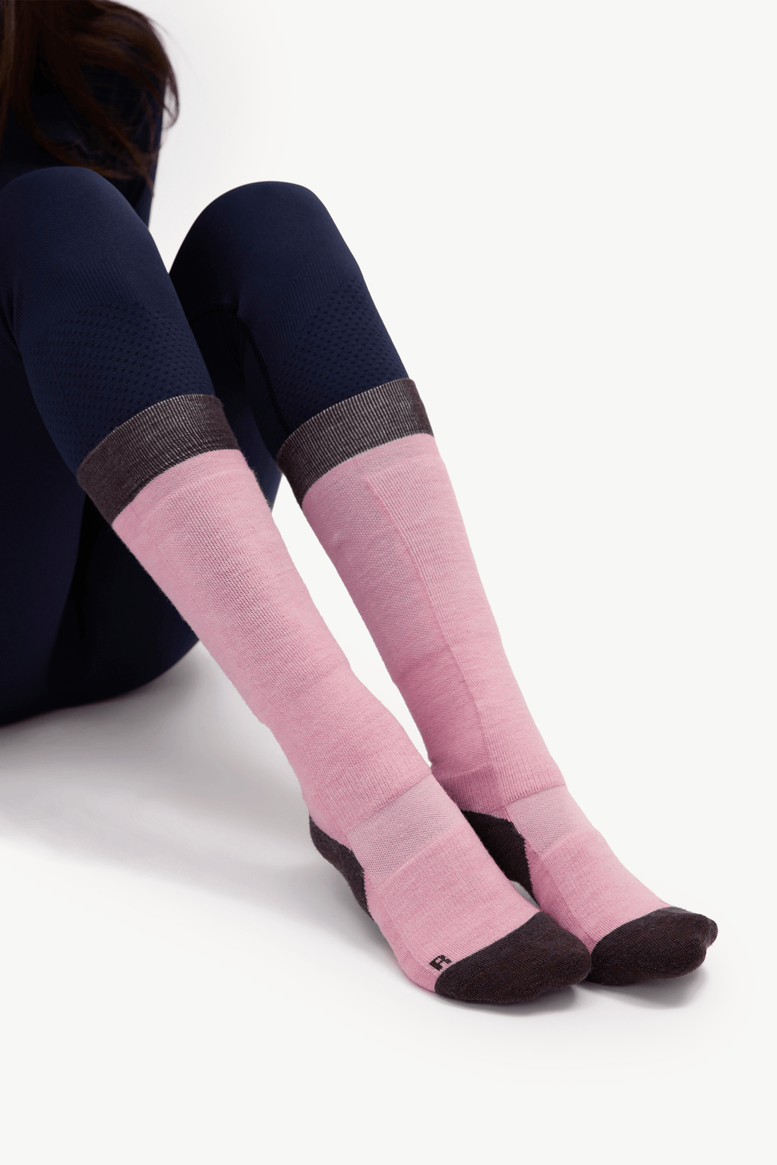 A person seated against a light gray studio background wearing knee-high pink socks with dark brown toes, heels, and soles, paired with dark navy leggings.